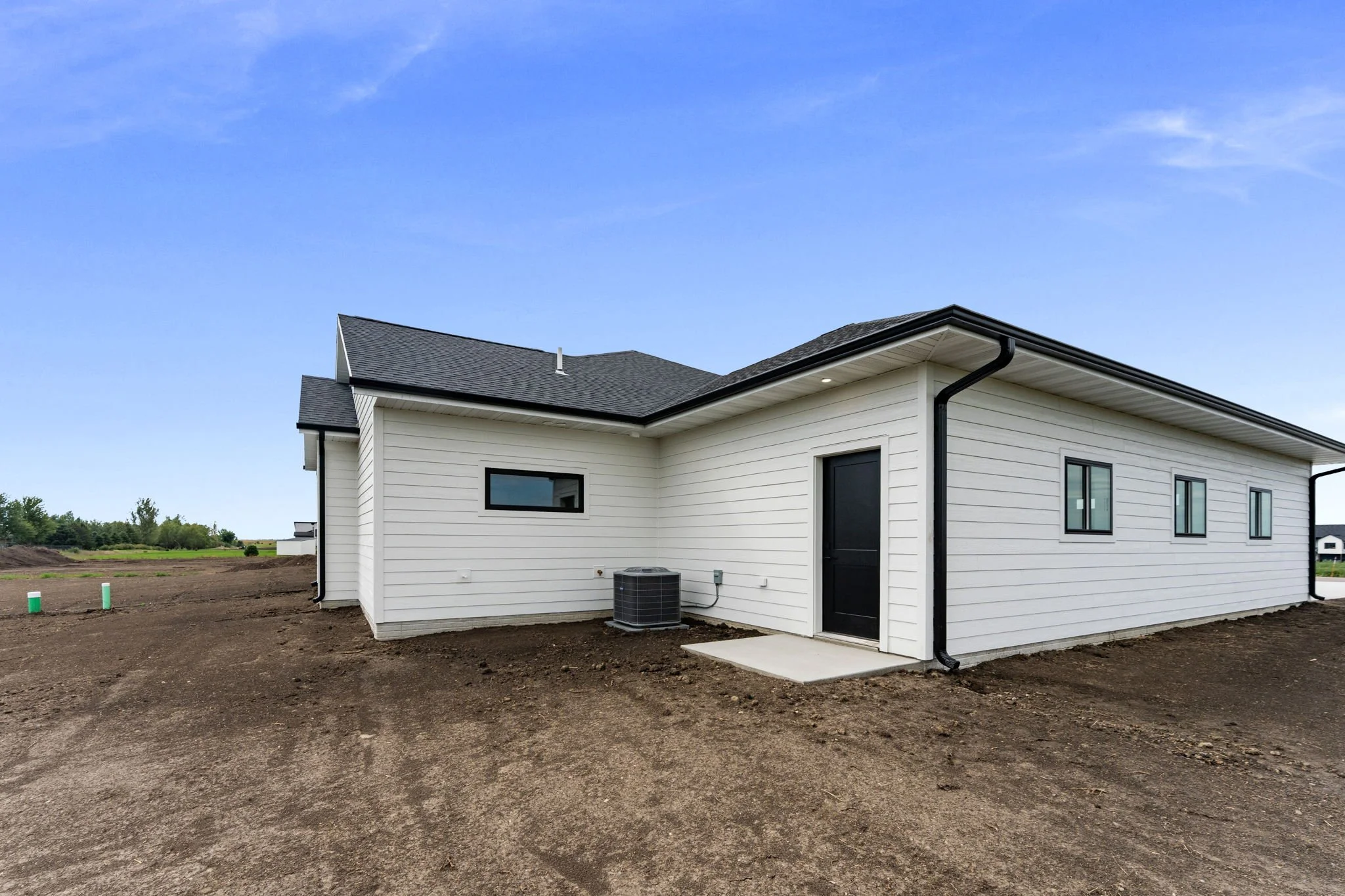 The image shows a modern, white, single-story house with a black door, black window frames, and a black roof. The house is situated in an open area with bare ground and distant trees and other homes in the background under a blue sky with some wispy 
