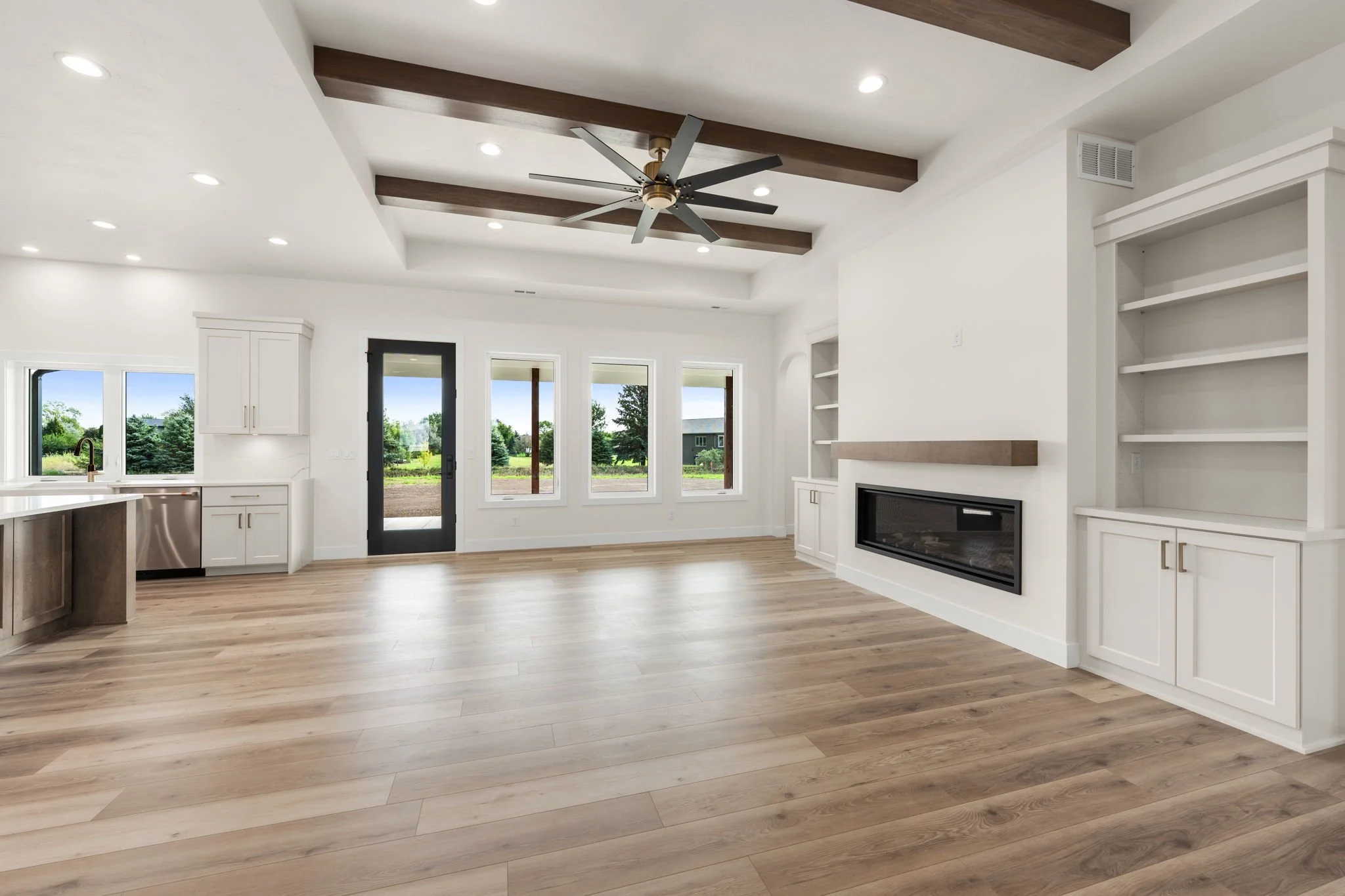 Empty living room with wood flooring, white walls, large windows, a fireplace, and a ceiling fan with dark blades and brass accents.