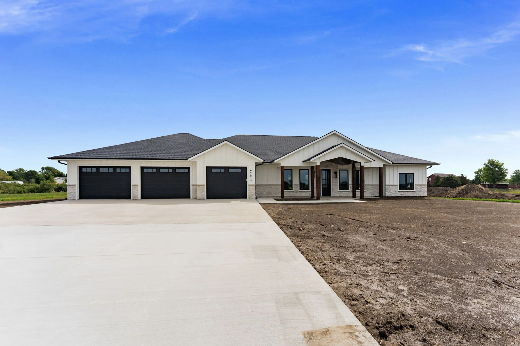A modern house with white siding and a black roof, featuring a three-car garage, a front porch supported by wooden columns, and a driveway, with a partly cloudy sky above and some grassy areas around.