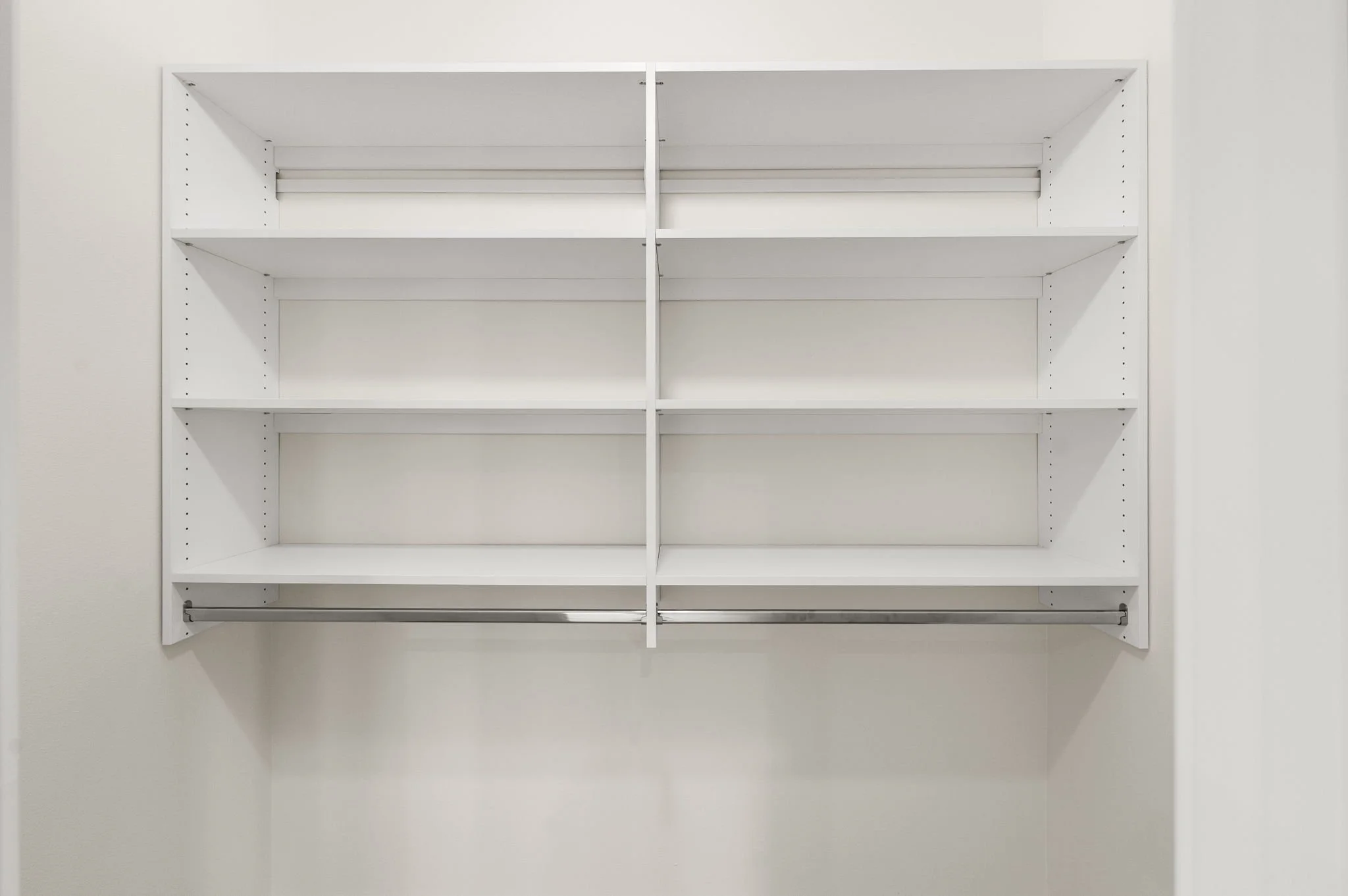 Empty white wall-mounted shelving unit with four tiers and a metal hanging rod at the bottom, against a plain white wall.