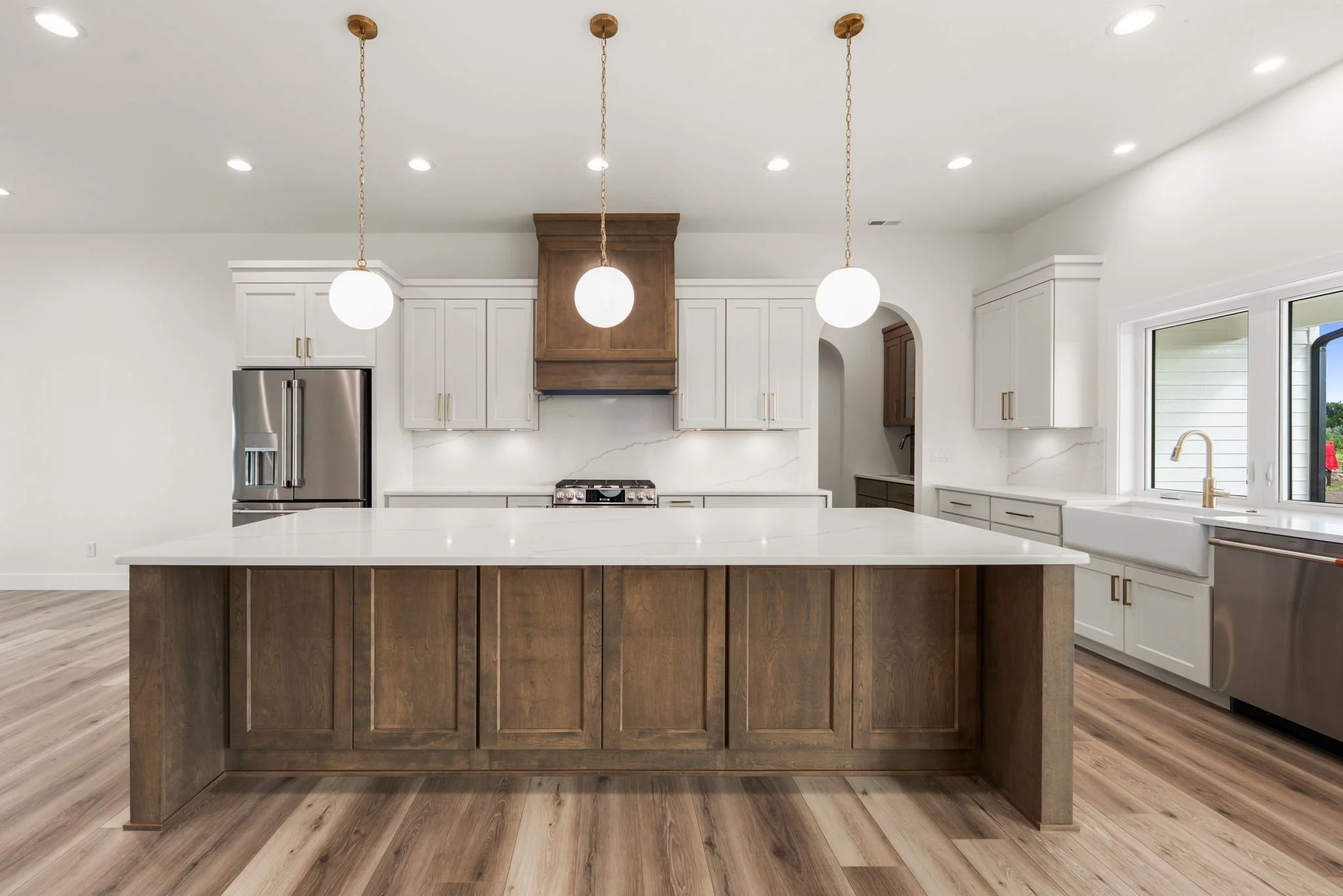 Modern kitchen with white cabinets, marble backsplash, stainless steel appliances, and a large island with a wooden base. Ceiling has three pendant lights and recessed lighting. Large window over the sink offers natural light.