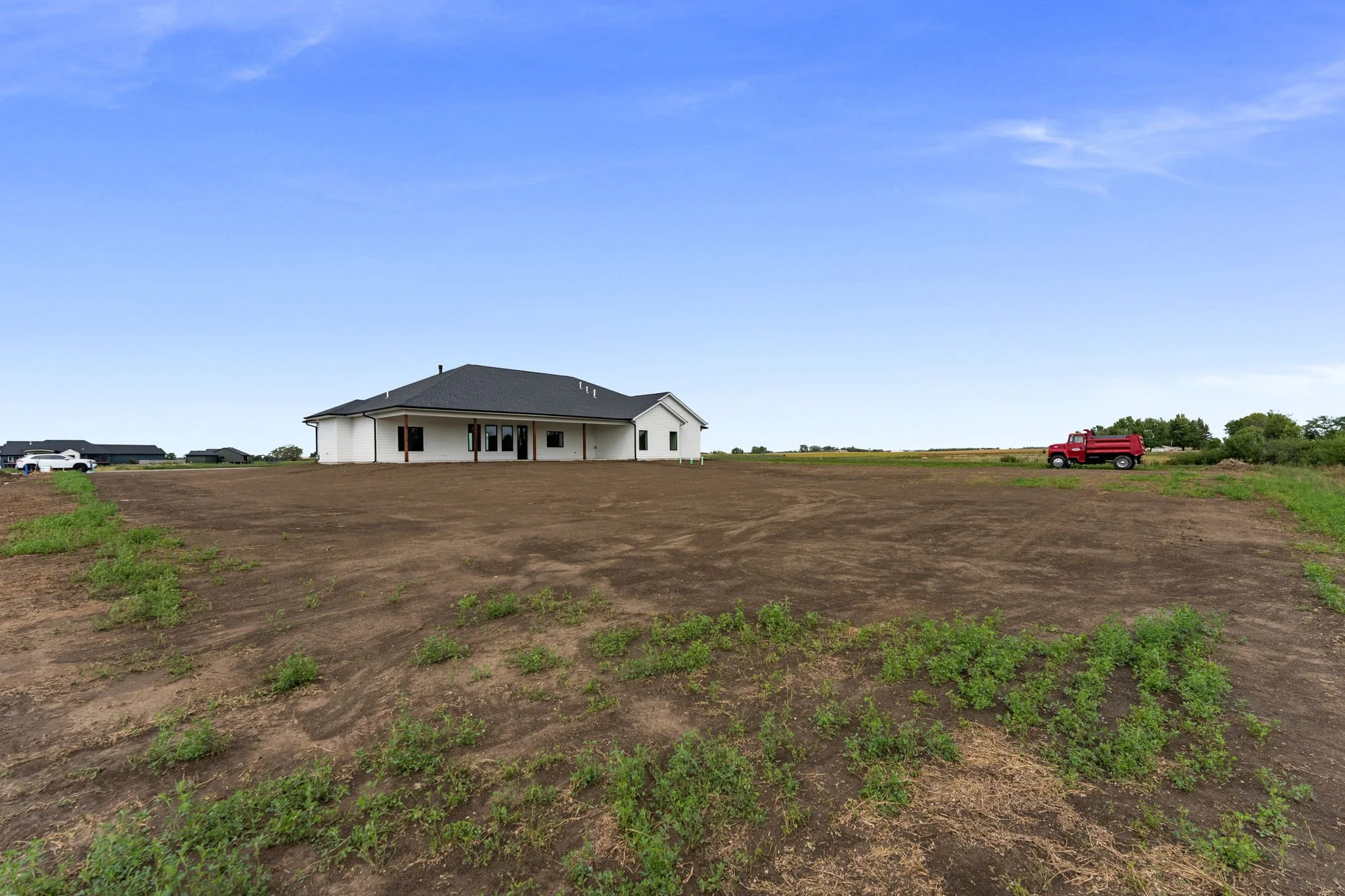 A house under construction on a large plot of land with dirt and few young plants, with a red truck parked to the right and other houses visible in the background under a partly cloudy blue sky.