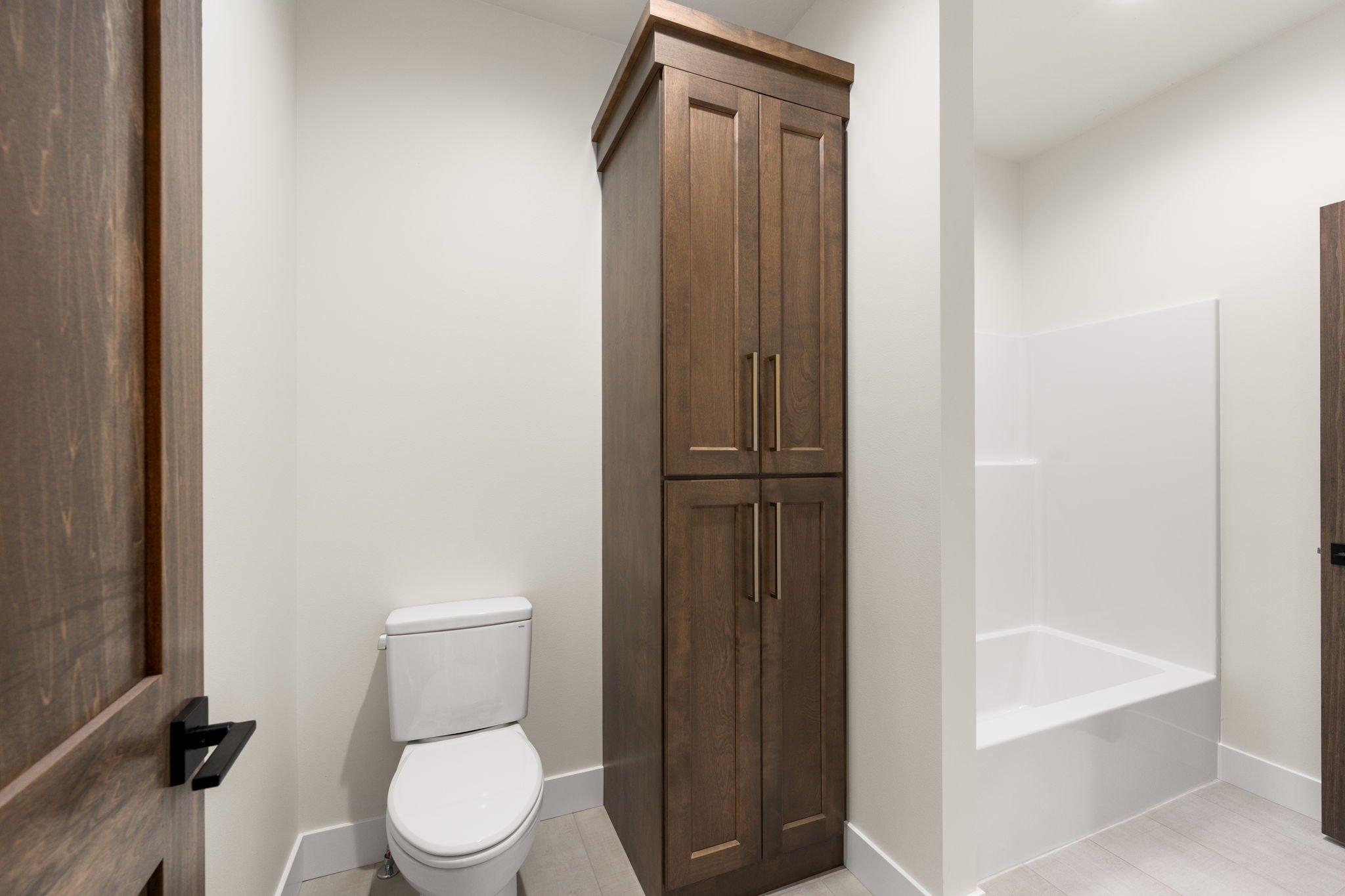 A modern bathroom with white walls and a white toilet. There is a tall wooden cabinet with doors and gold handles next to the toilet. A portion of a white bathtub with a shower is visible to the right. The bathroom has a minimalist decor with clean l