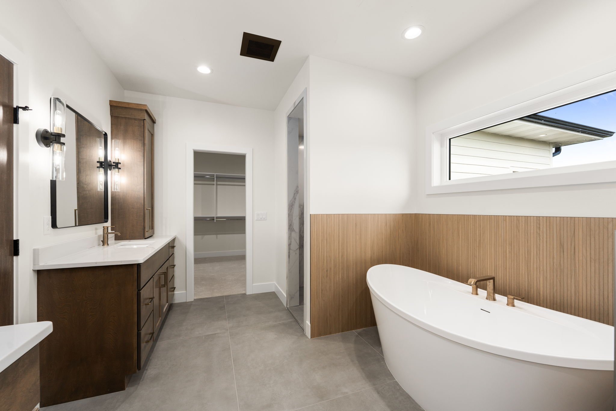 Modern bathroom with a white bathtub, wood paneling, white walls, a vanity with dark wood cabinets, a large mirror, and a walk-in closet visible in the background. There's a window on the right side and a ceiling vent.