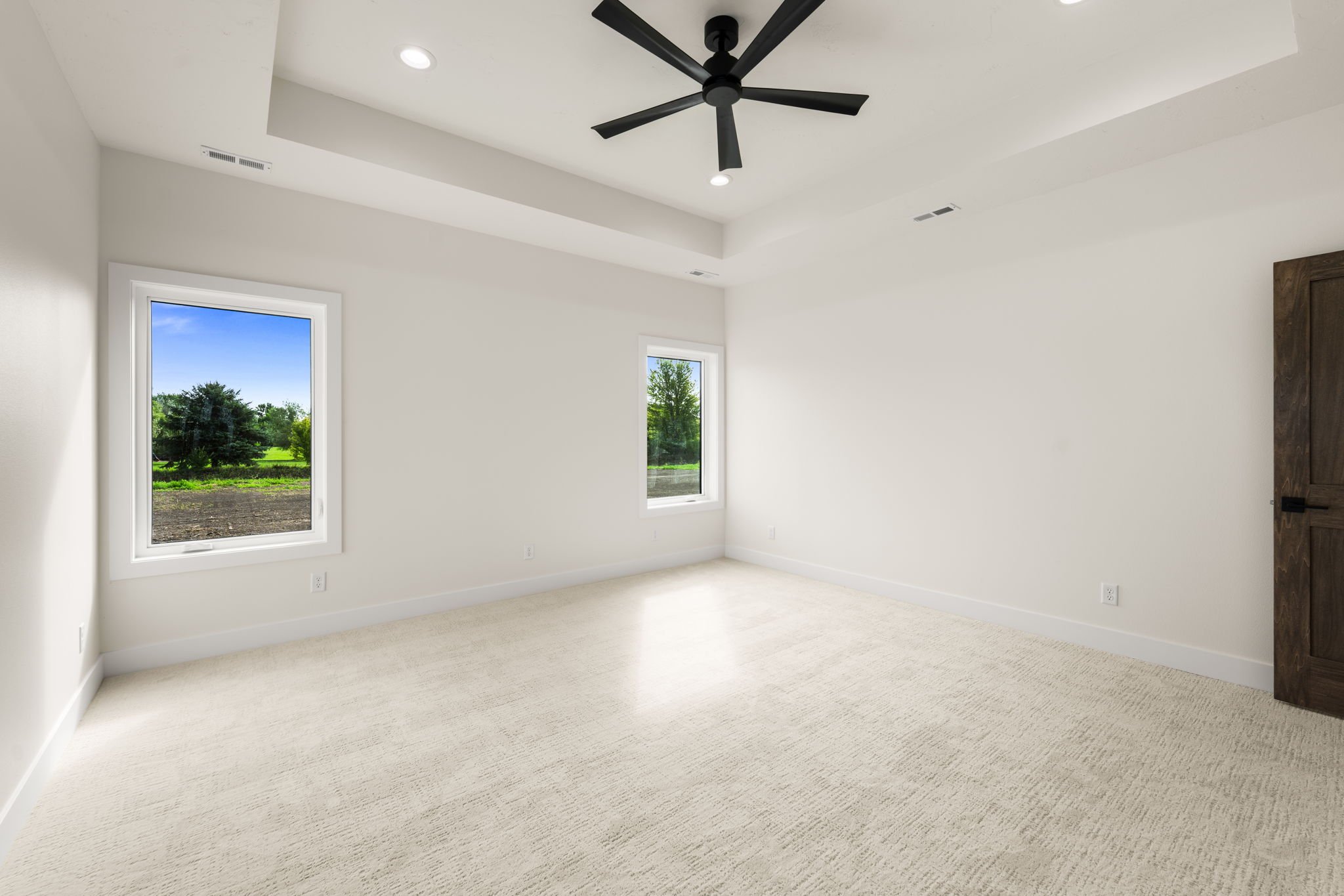 An empty, bright room with white walls, beige carpet, two windows showing green trees and grass outside, a black ceiling fan, and a wooden door.