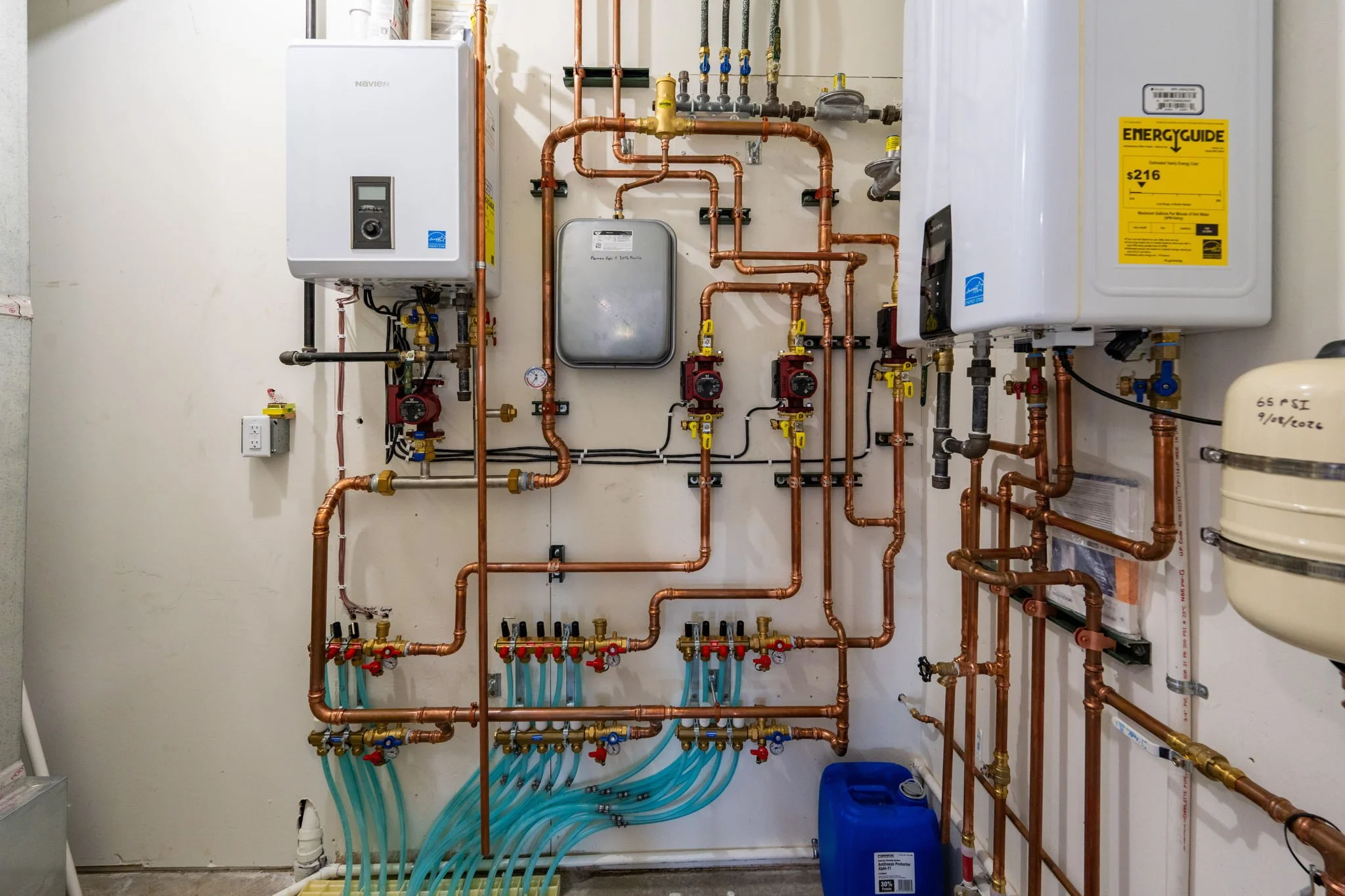A utility room with various pipes, gauges, and water meters installed on a wall, including a water heater, a pressure tank, and multiple copper pipes with valves.