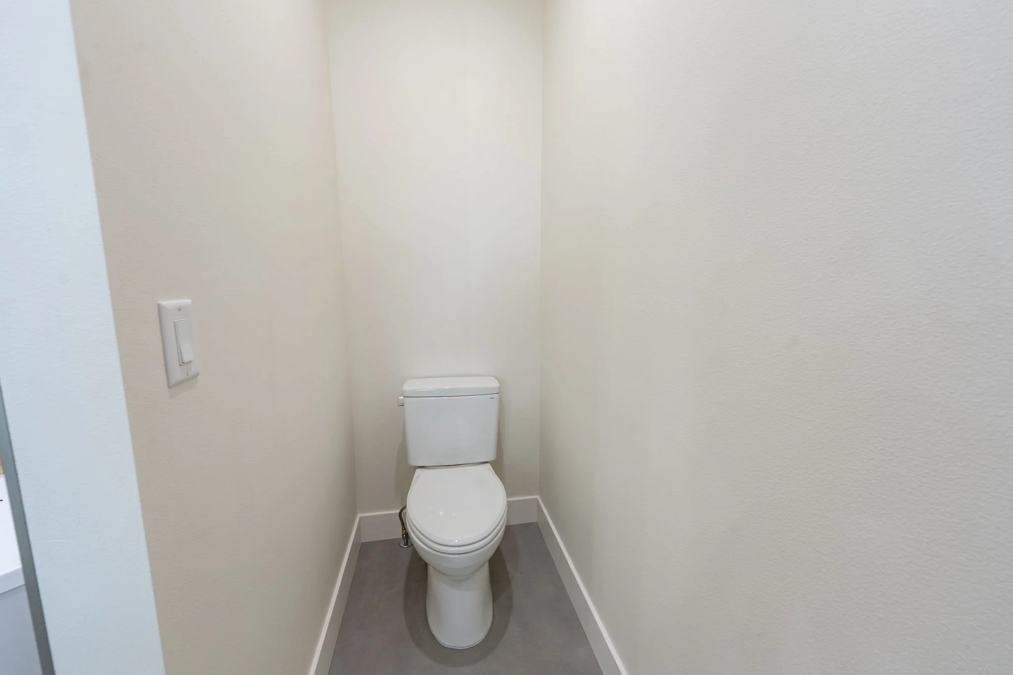 A small, plain bathroom with beige walls, a white toilet, a light switch on the wall, and gray flooring.