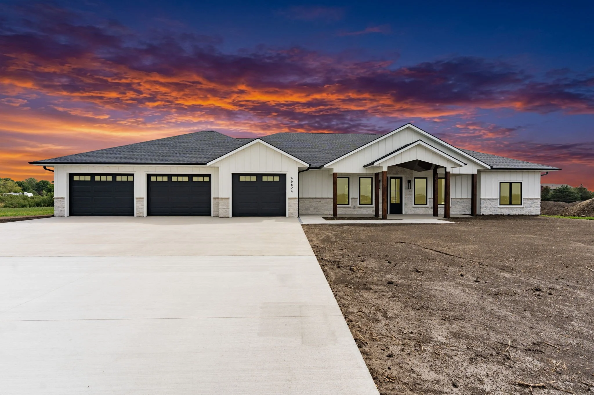A newly constructed house with a dark gray roof, white siding, and a four-car garage, with a sunset sky in the background and a concrete driveway leading up to it.