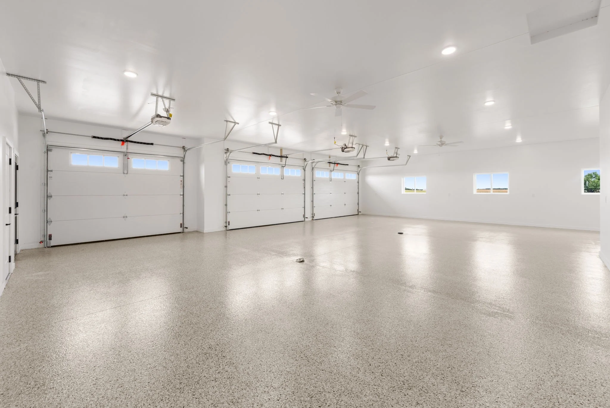 A spacious clean garage with white walls, ceiling, and garage doors, equipped with ceiling fans and bright overhead lights.