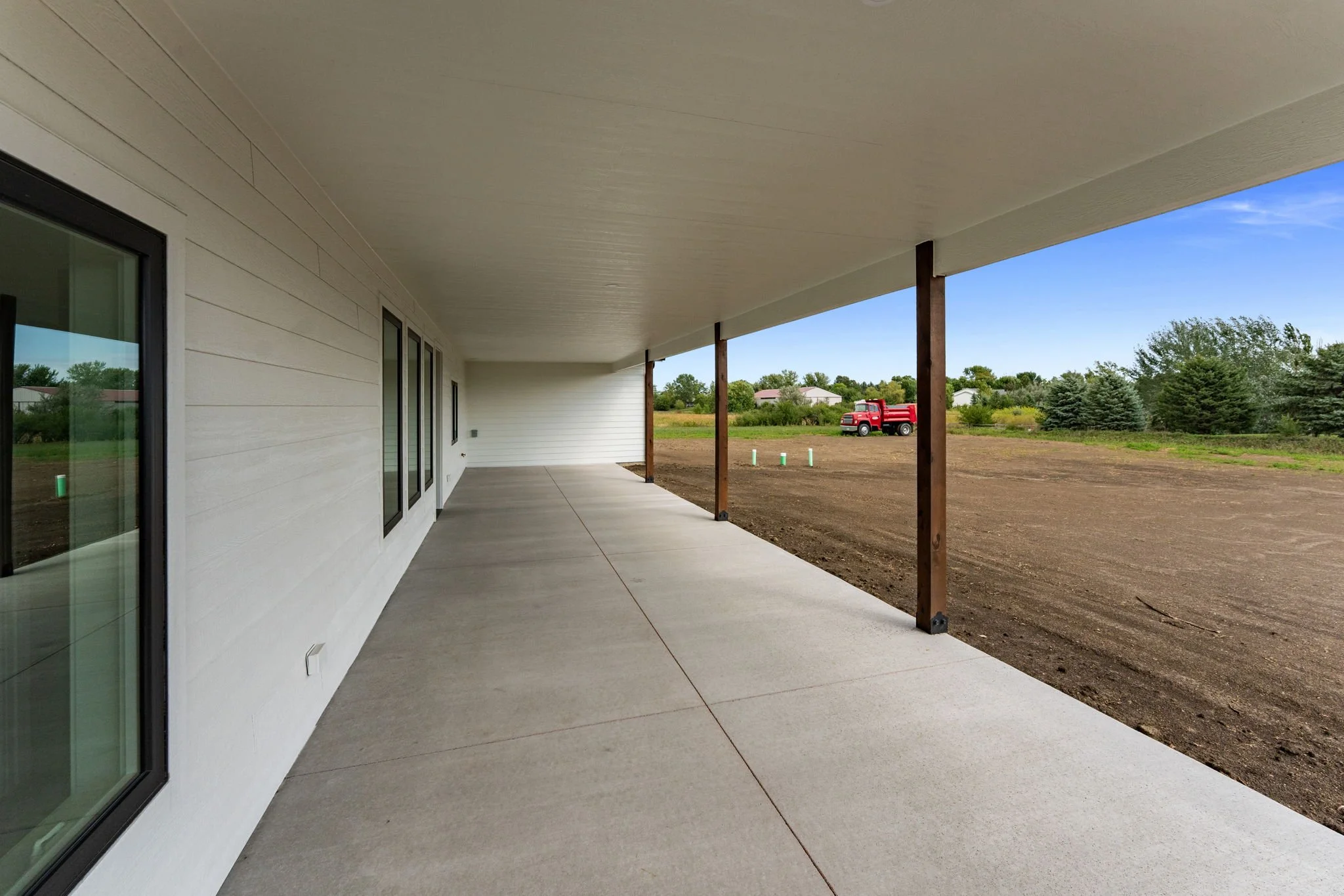 Newly built house with a covered porch, concrete flooring, beige exterior walls, and large glass windows. There are wooden support posts on the porch, and in the background, there is a dirt yard with a red truck and some greenery.