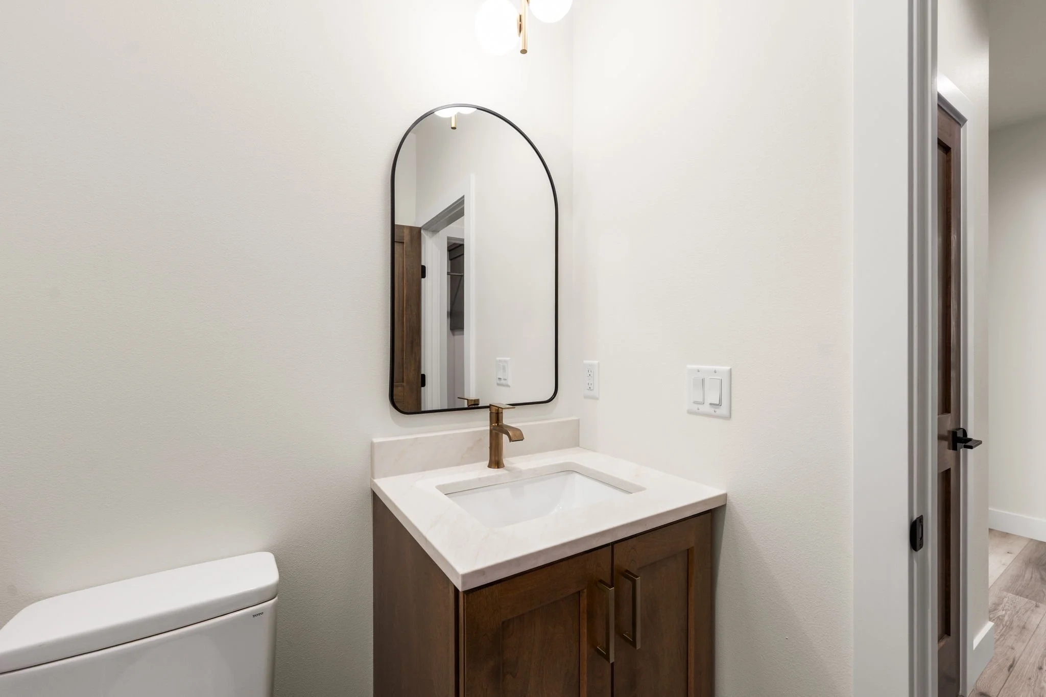 Bathroom vanity with a large oval mirror, gold faucet, and wooden cabinet, with a toilet partially visible on the left and a doorway on the right.
