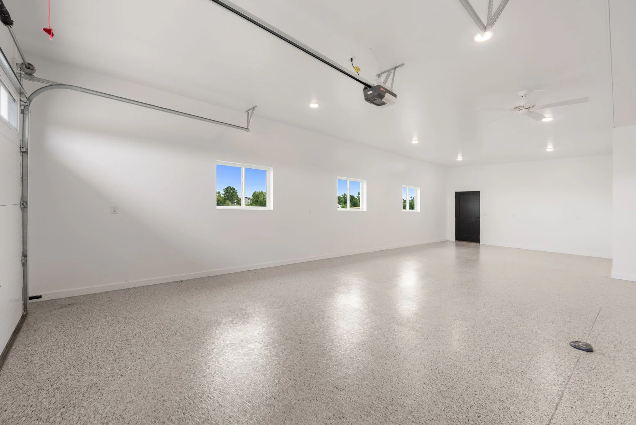 Empty residential garage with white walls, a concrete floor, three small windows, a black door, ceiling lights, and a garage door opener.