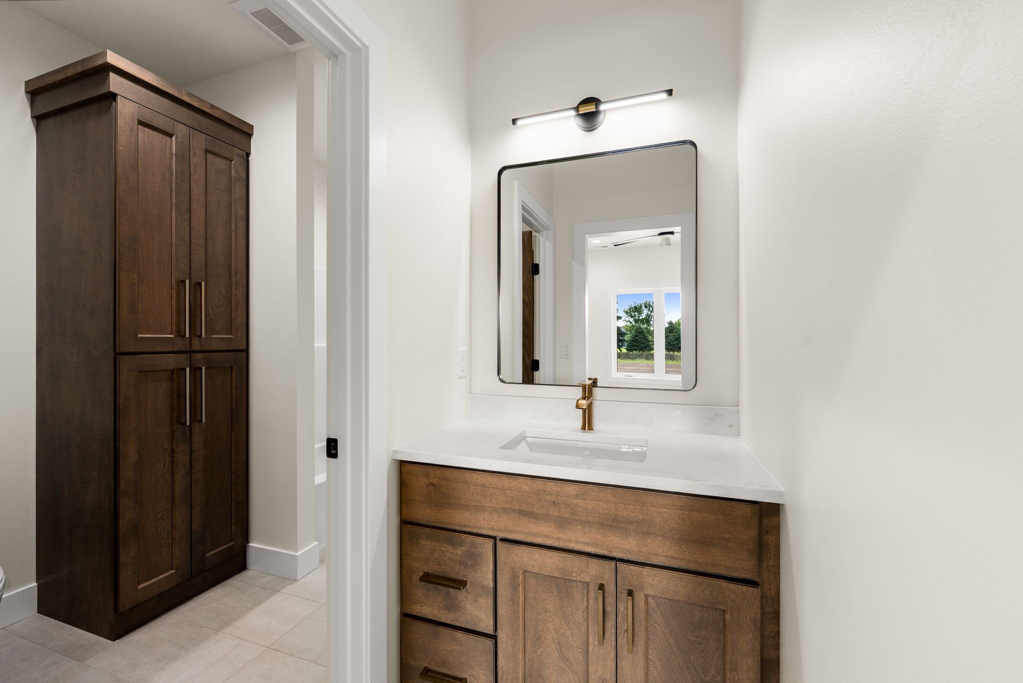 Modern bathroom with wooden cabinets, a white marble countertop, a rectangular mirror, and a window showing a view of trees outside.