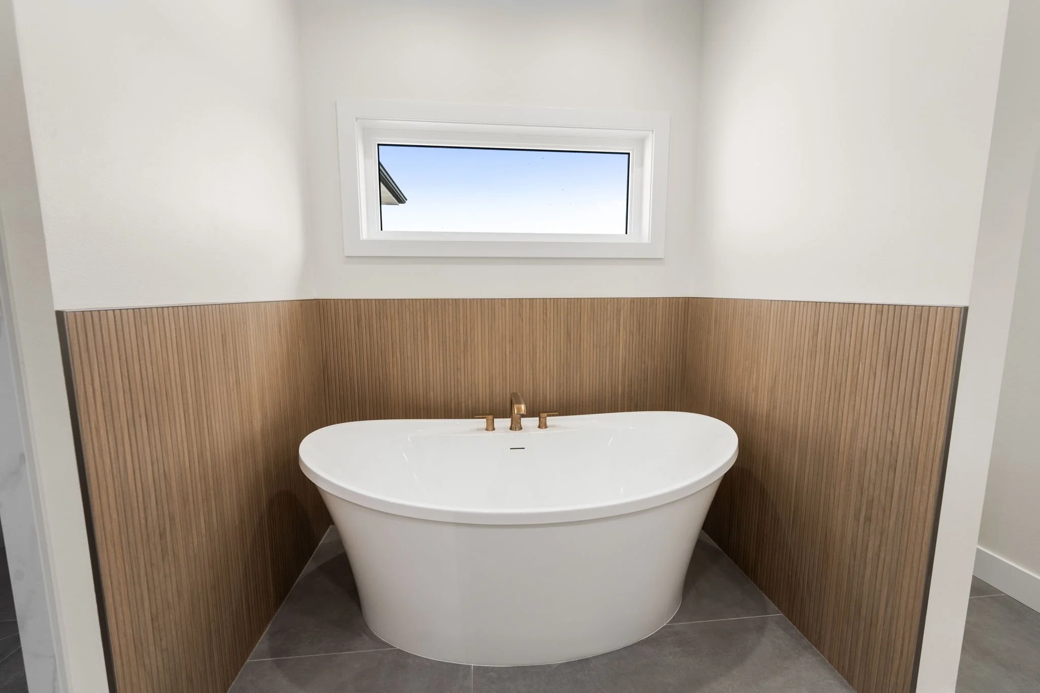 A modern bathroom with a freestanding bathtub under a rectangular window, with wooden paneling on the walls and gray tile flooring.