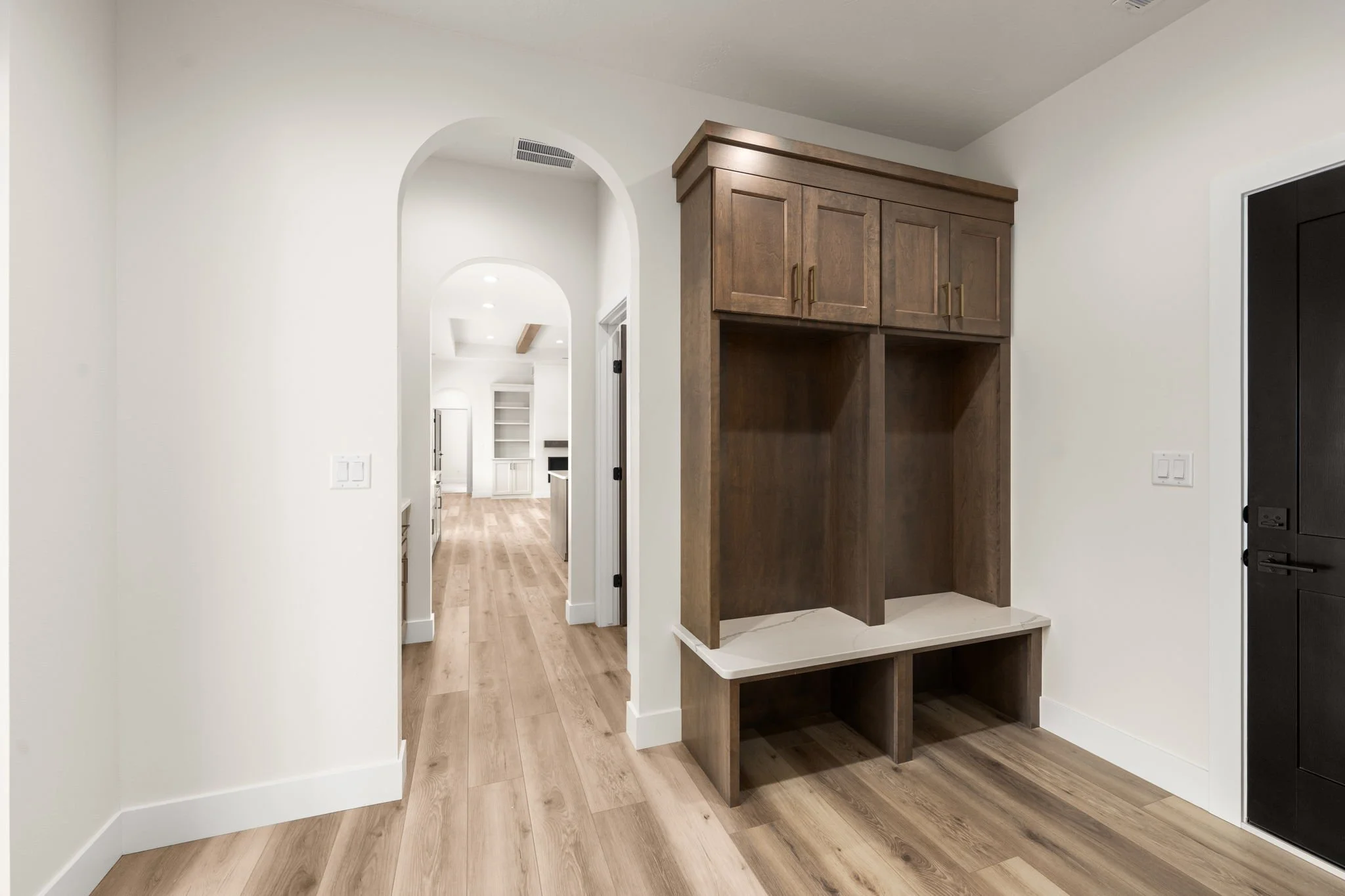 Interior of a modern home with wood flooring, white walls, arched doorways, and built-in cabinets in dark wood. The hallway leads to other rooms, and a black door is visible on the right.
