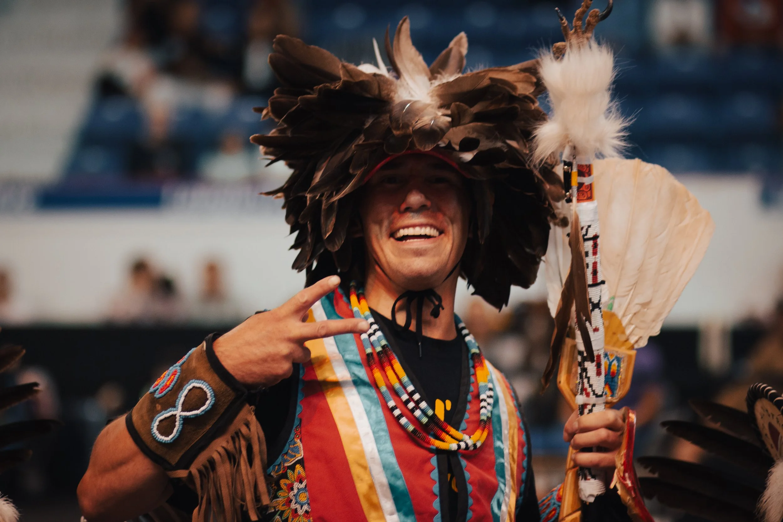 A man smiling and making a peace sign with his right hand, wearing traditional Native American attire including a large feathered headdress and beaded necklaces at a cultural event or celebration.