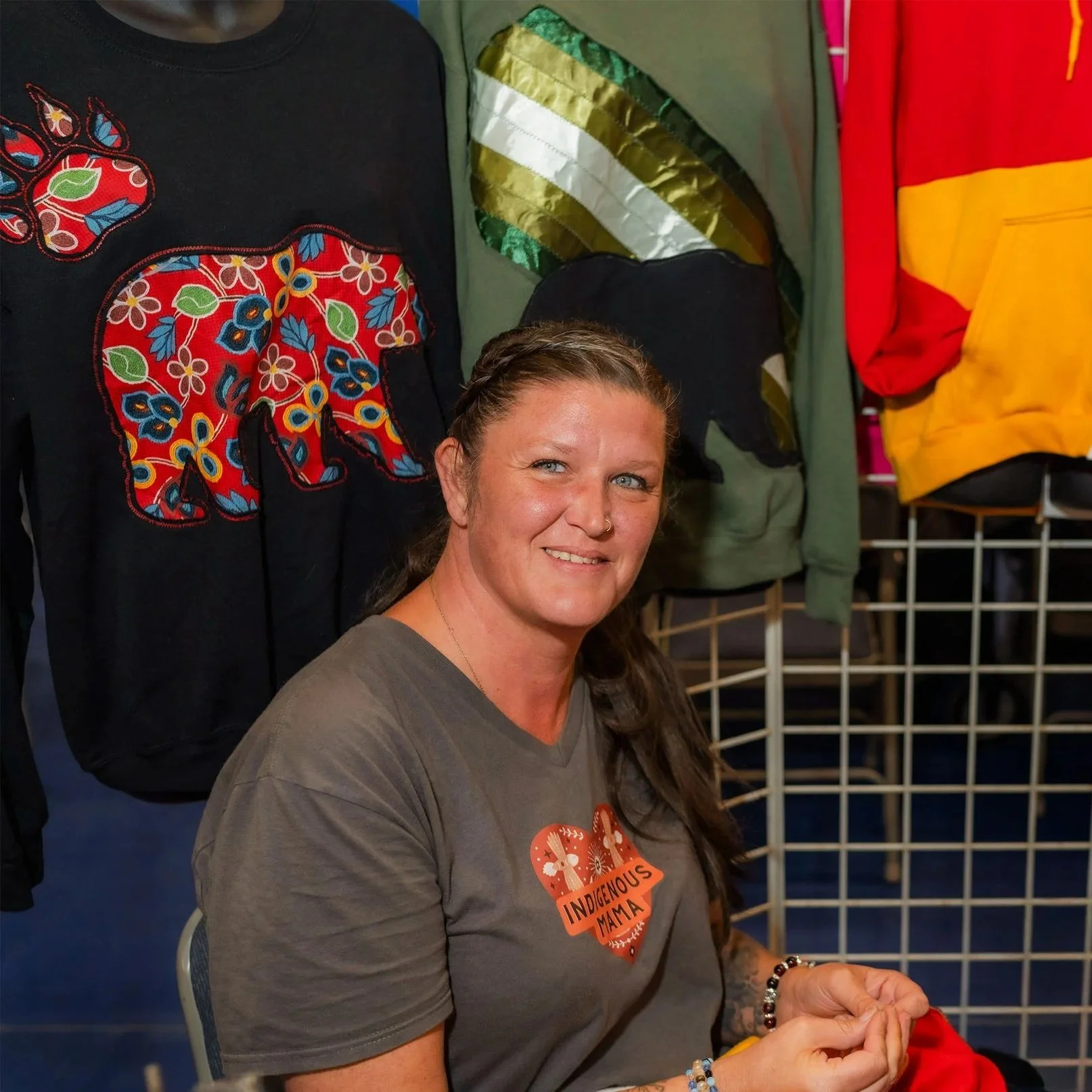 A woman with long brown hair and a nose piercing smiling while sitting at a booth with colorful jackets and sweatshirts behind her, one with a floral pattern in a bear shape, and wearing a gray T-shirt with an 'Indigenous Mama' sticker.