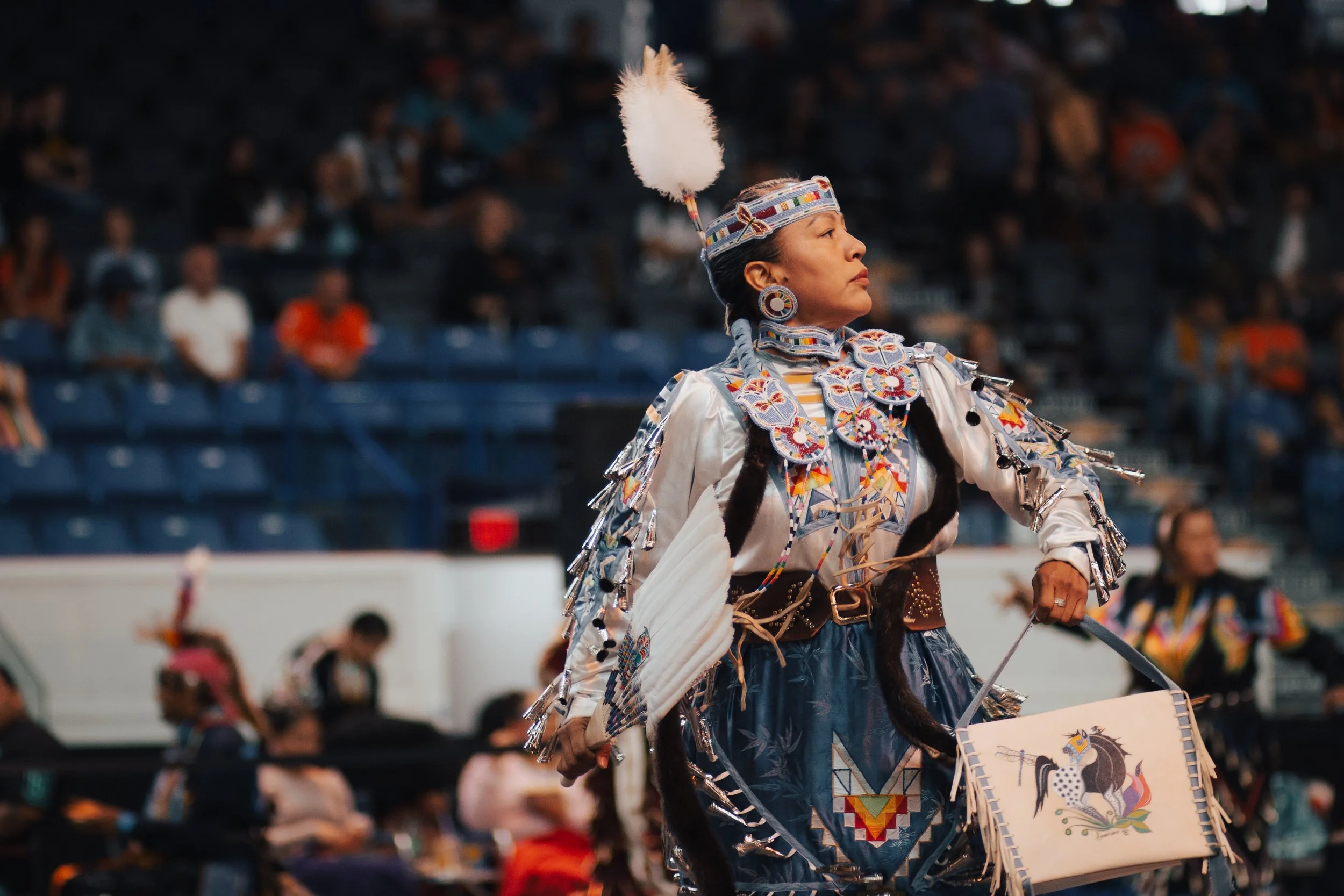A Native American woman in traditional dress, decorated with beads, fringes, and a feather headdress, holding a blanket with a horse design, participating in a cultural event or ceremony.