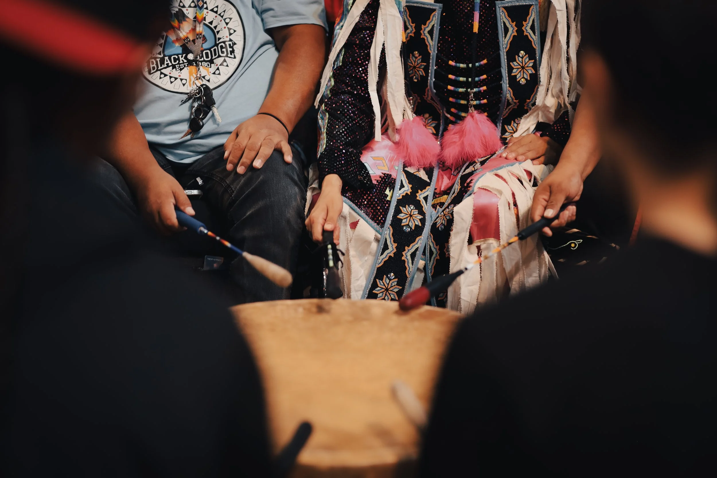 People sitting around a wooden drum, holding drumsticks, with one person dressed in traditional Native American attire.