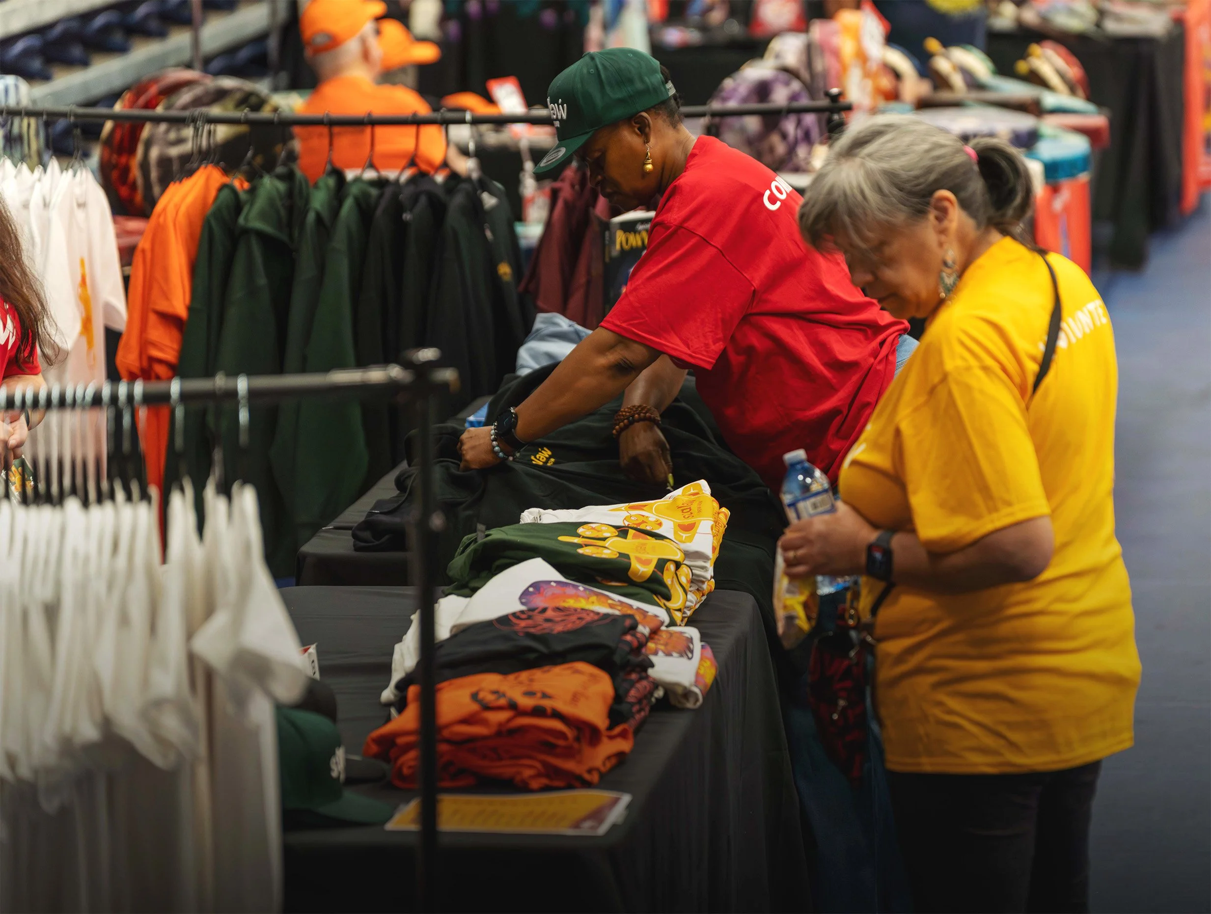 Two women browsing merchandise at a table in a retail store, with clothing and hats displayed around them.