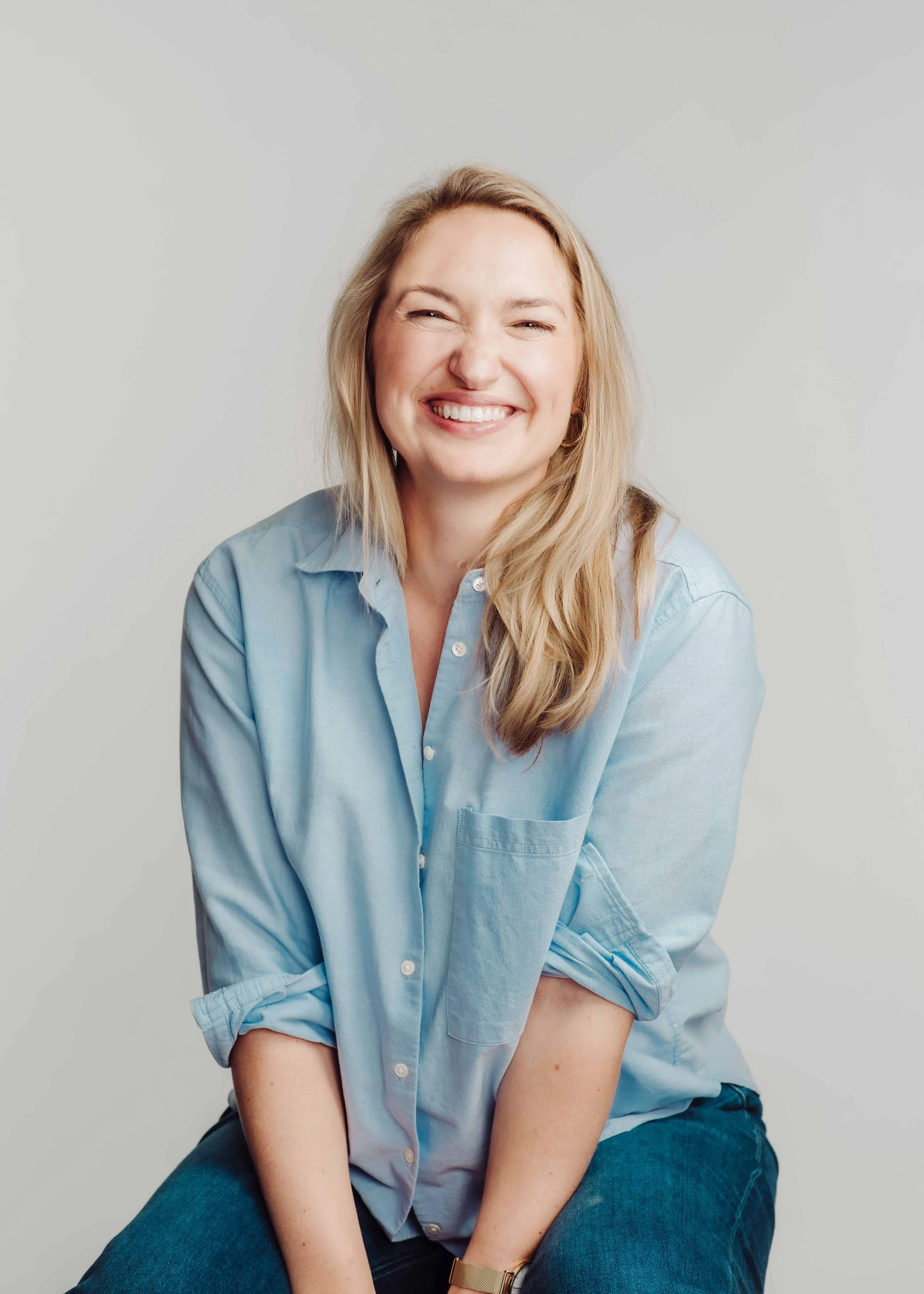 A smiling woman with blonde hair, wearing a light blue button-up shirt and blue jeans, sitting against a plain light gray background.