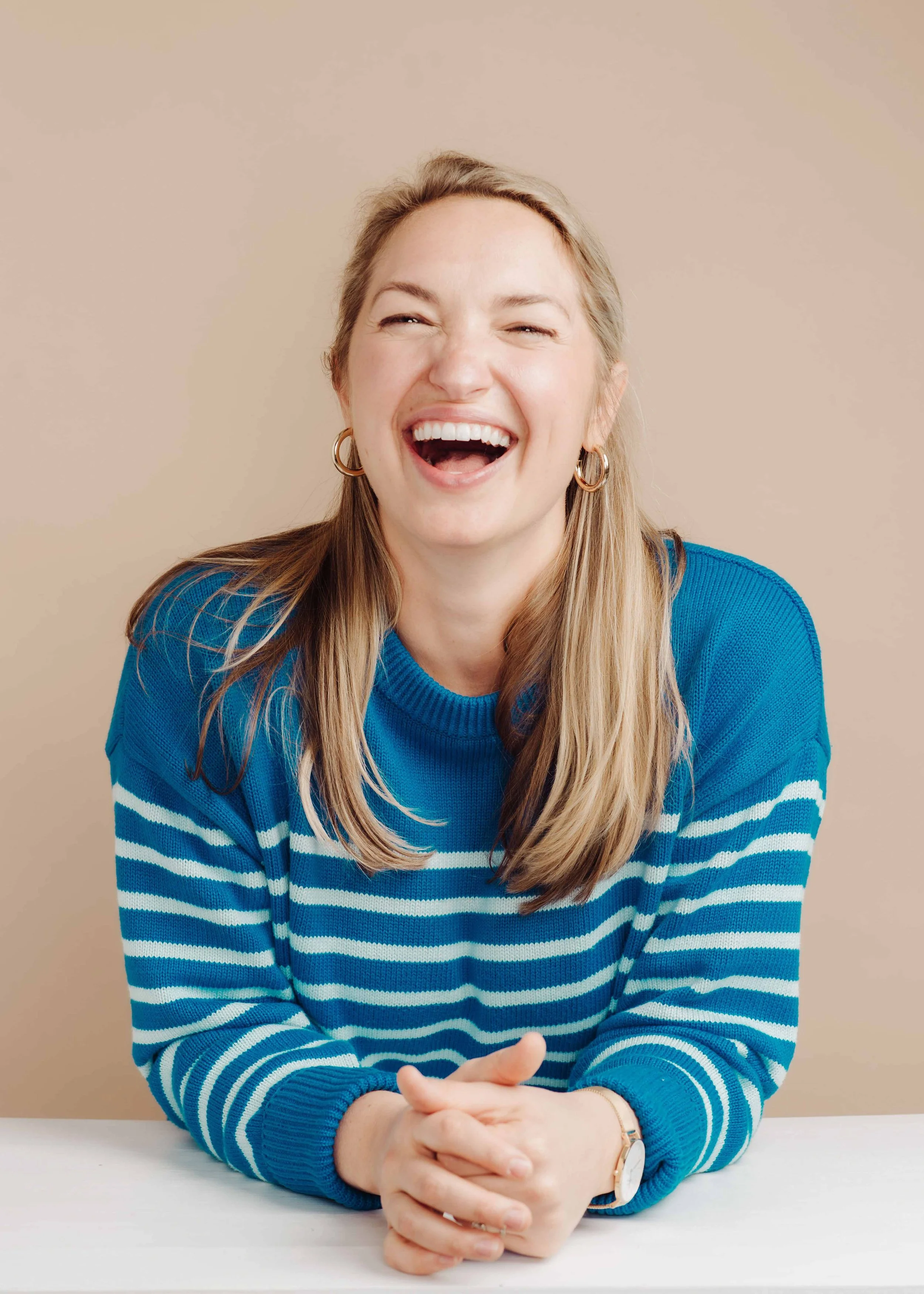 A woman with long blonde hair, wearing a blue and white striped sweater, sitting at a white table, laughing with her eyes closed and hands clasped.
