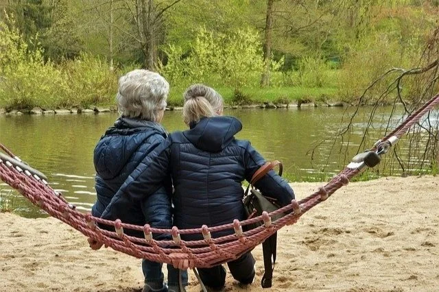Two elderly women sitting on a hammock near a lake, surrounded by trees, enjoying the outdoors.