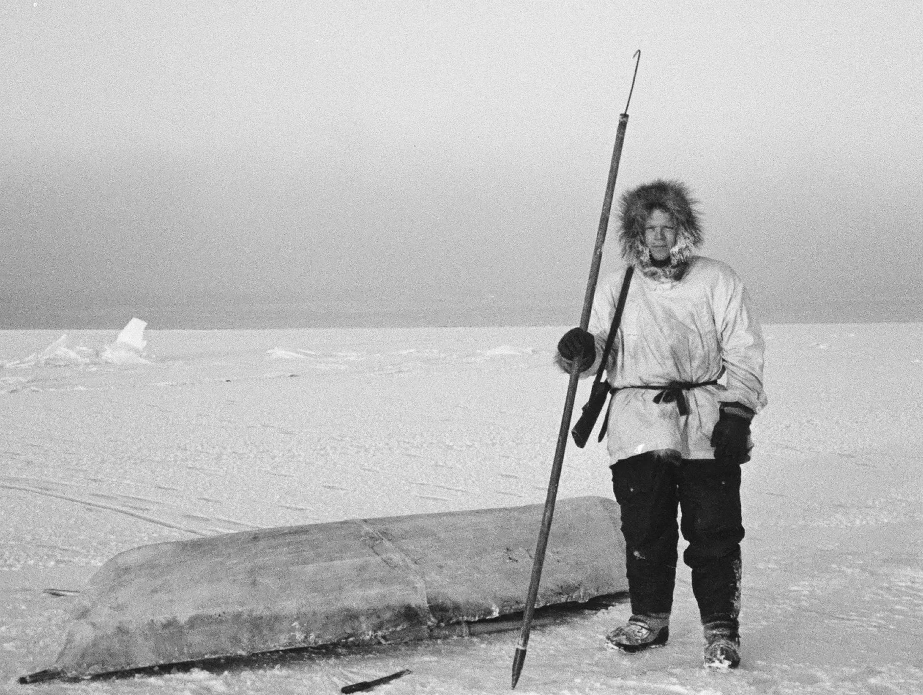Richard Nelson on young ice with open water in the background. Rifle over his shoulder, he holds an unaaq used to test ice thickness. The umiaqaluraq is used to retrieve seals. (Wainwright, Alaska 1964)