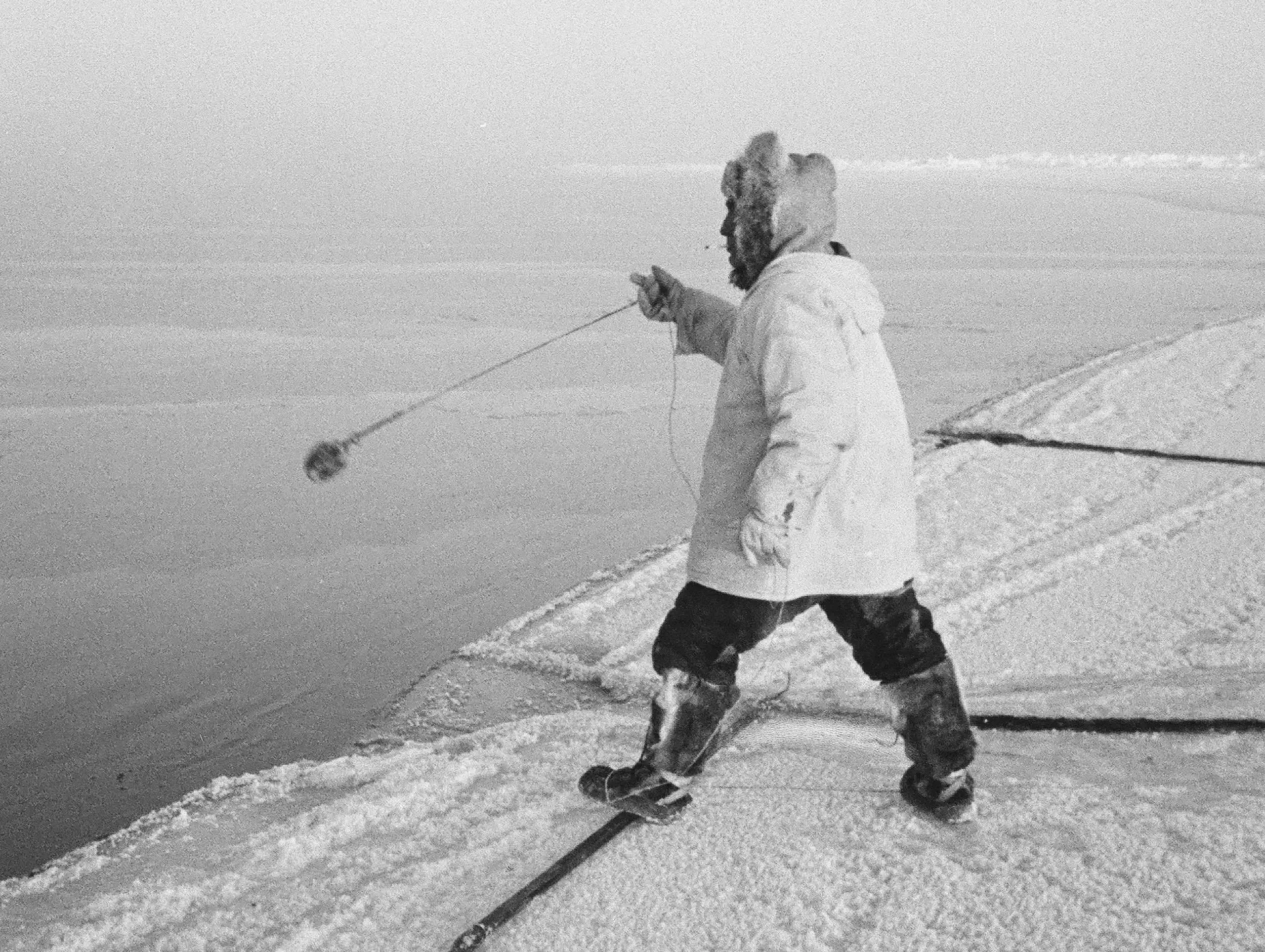 Anaqqaq Ekak tossing a manaq (wooden float with metal hooks) to retrieve a seal shot in open water. He's standing on his unaaq to distribute his weight on the thin ice.