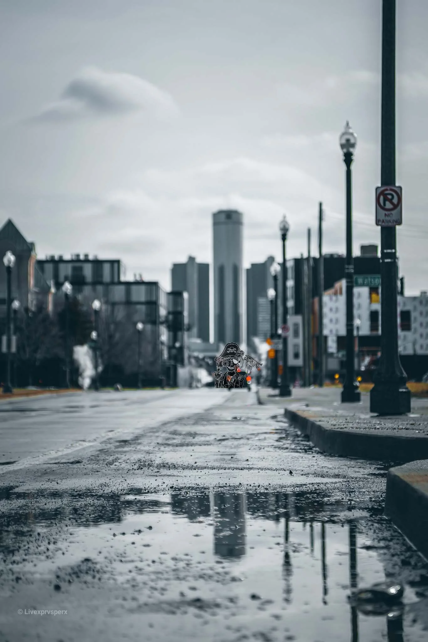 City street scene with puddles on the road, tall buildings in the background, and streetlamps. A person with an umbrella is walking down the sidewalk.