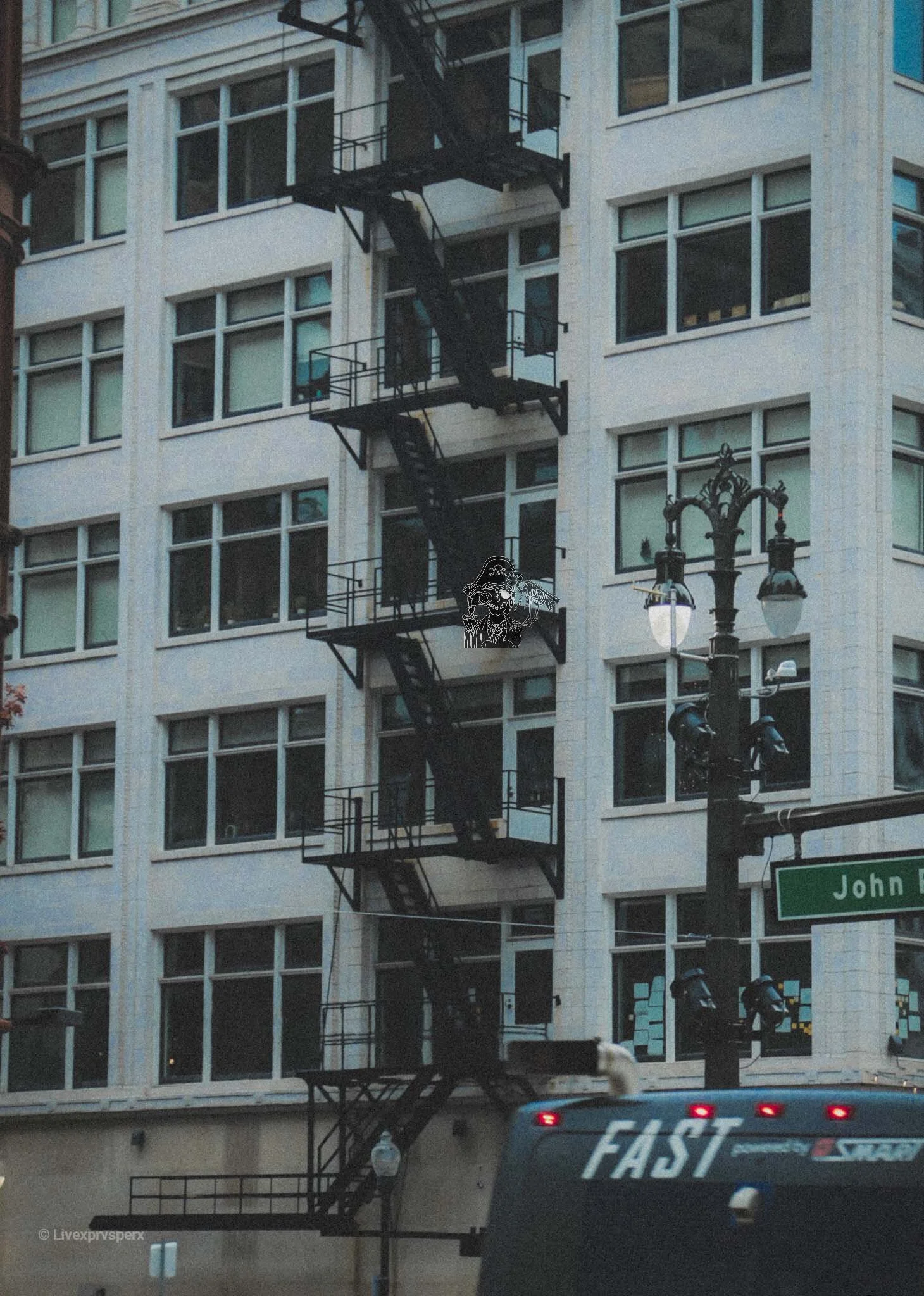 A city street scene with a tall building, fire escape stairs on the building's facade, a street lamp with two lights, and a vehicle with the word 'FAST' on it passing by. A street sign reading 'John' is partially visible.