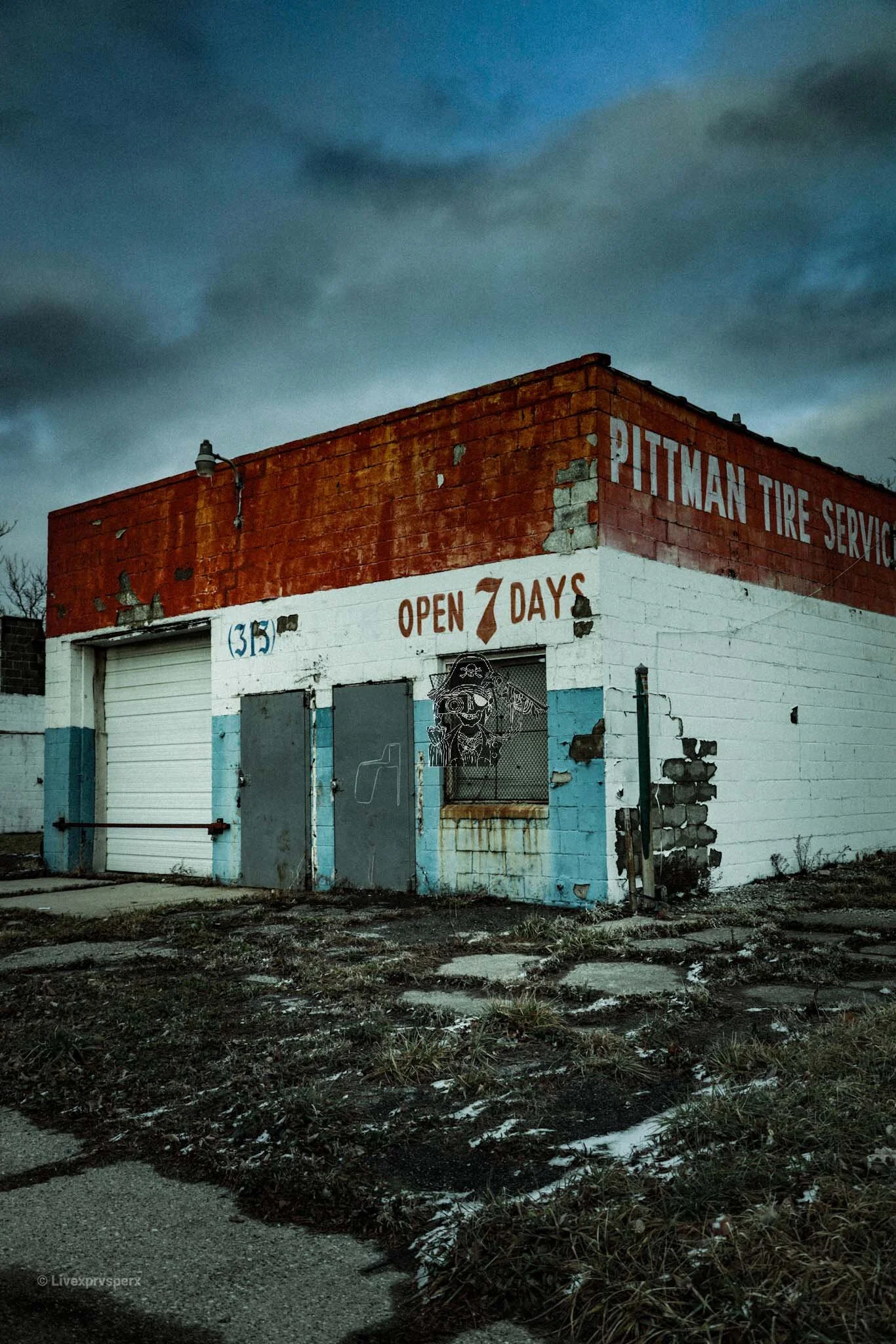 A worn-out brick and concrete building of Pittman Tire Service with an open 7 days sign, graffiti art, and a dark, cloudy evening sky.