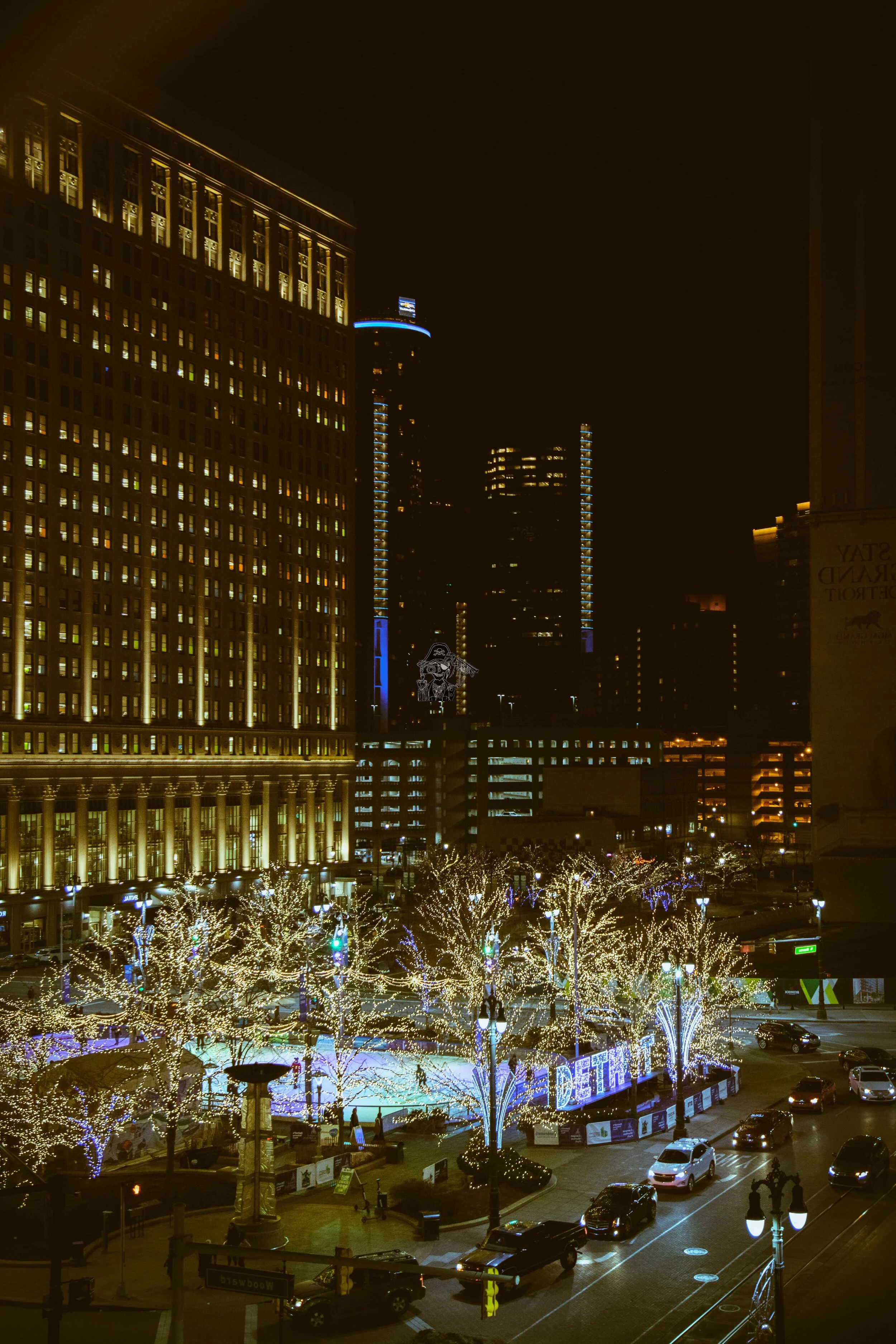 Nighttime city scene with illuminated trees surrounding a skating rink, tall buildings with lights, and traffic on the street.
