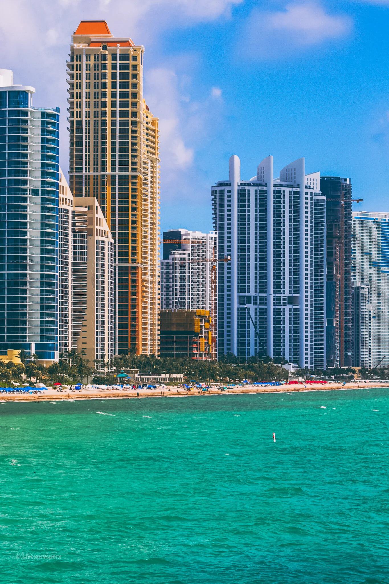 City skyline with tall modern skyscrapers along a beach, turquoise water, and blue sky with scattered clouds.