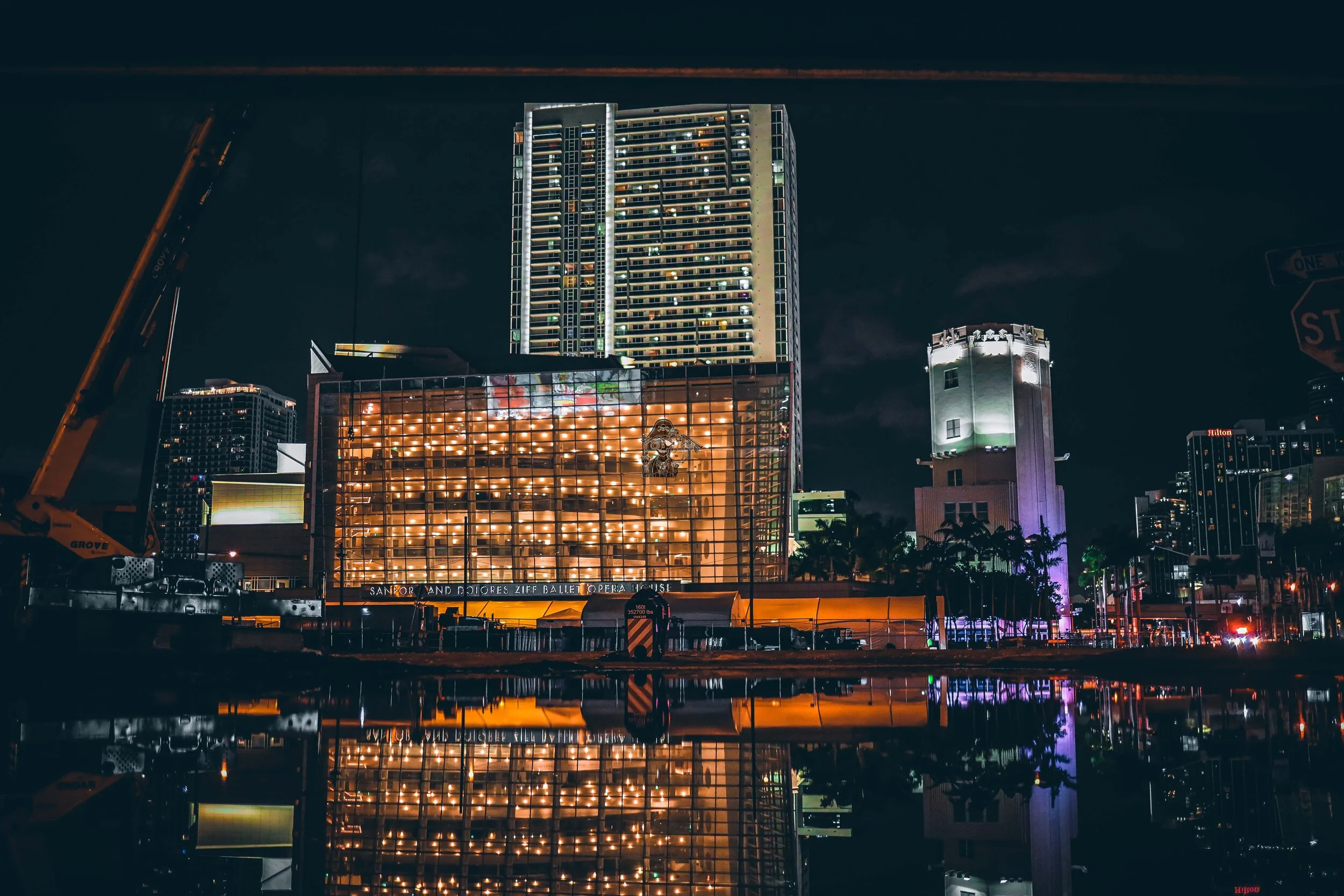 Nighttime cityscape with illuminated modern buildings, a tall skyscraper, and reflections on water in the foreground.
