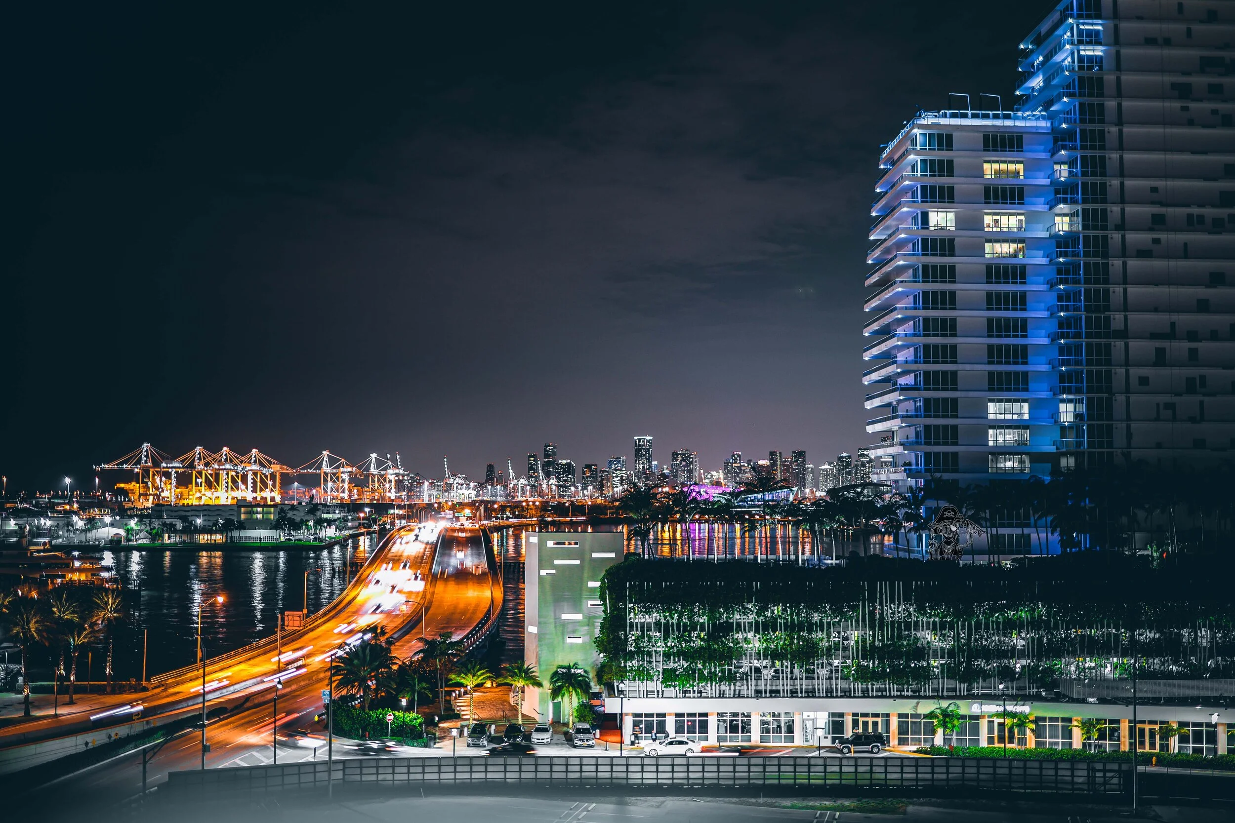 Nighttime cityscape with illuminated buildings, a bridge with bright lights, a highway with moving headlights and taillights, and a high-rise building with blue lighting on one side.