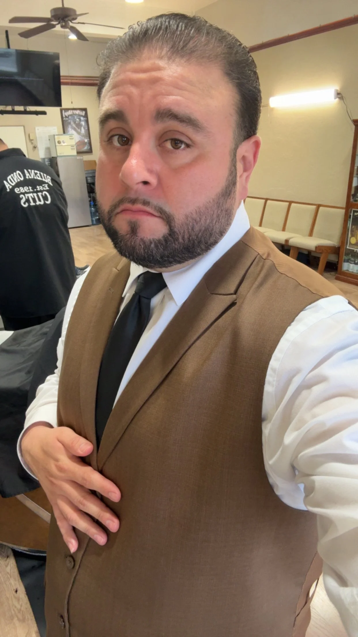 Owner of Privileged Professional Barbershoo Juan ”Kookie" is A man with a beard and slicked-back hair wearing a brown vest, white shirt, and black tie taking a selfie in a room with beige walls, wooden furniture, and a ceiling fan.
