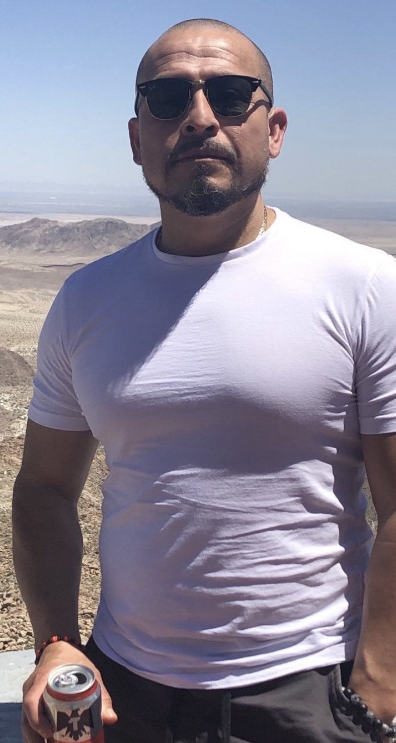 Rafa is a Barber at Privileged Professional Barbershop and is the man wearing sunglasses and a white t-shirt standing outdoors in a desert landscape, holding a can of soda.