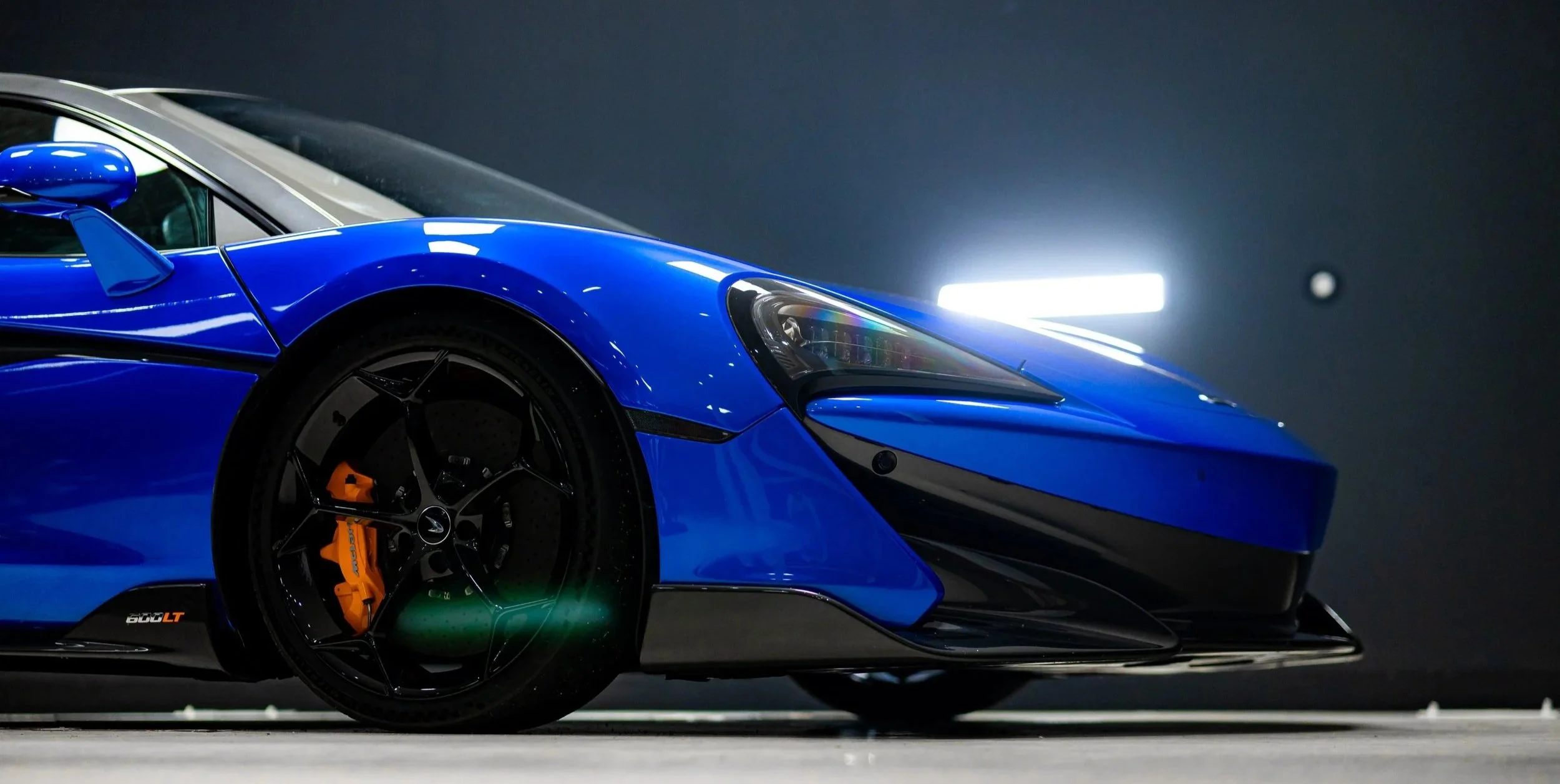 Close-up of a blue sports car, showing the front left side, wheel with black rim and orange brake caliper, sleek headlight, and aerodynamic design in a dark indoor setting with bright lights in the background.