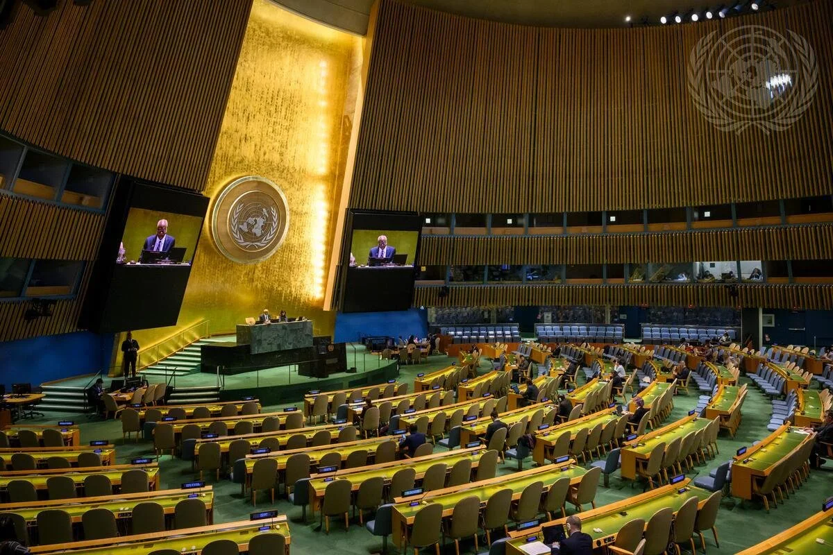 United Nations General Assembly hall with rows of yellow seats, a large globe emblem on a golden wall behind the speaker's podium, and a large screen displaying a speaker.