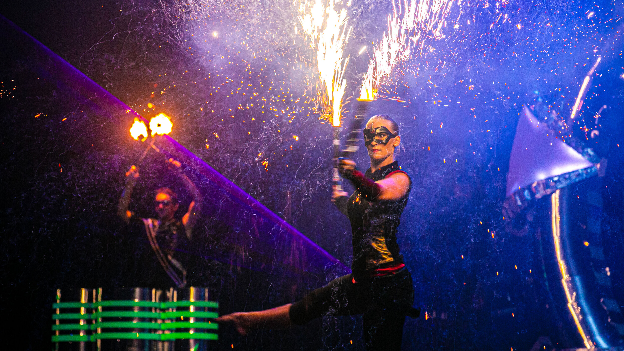 Elegante Tänzerin mit Pyro Excess Effekt in Händen und im Hintergrund Feuertrommler mit brennenden Trommelstöcken