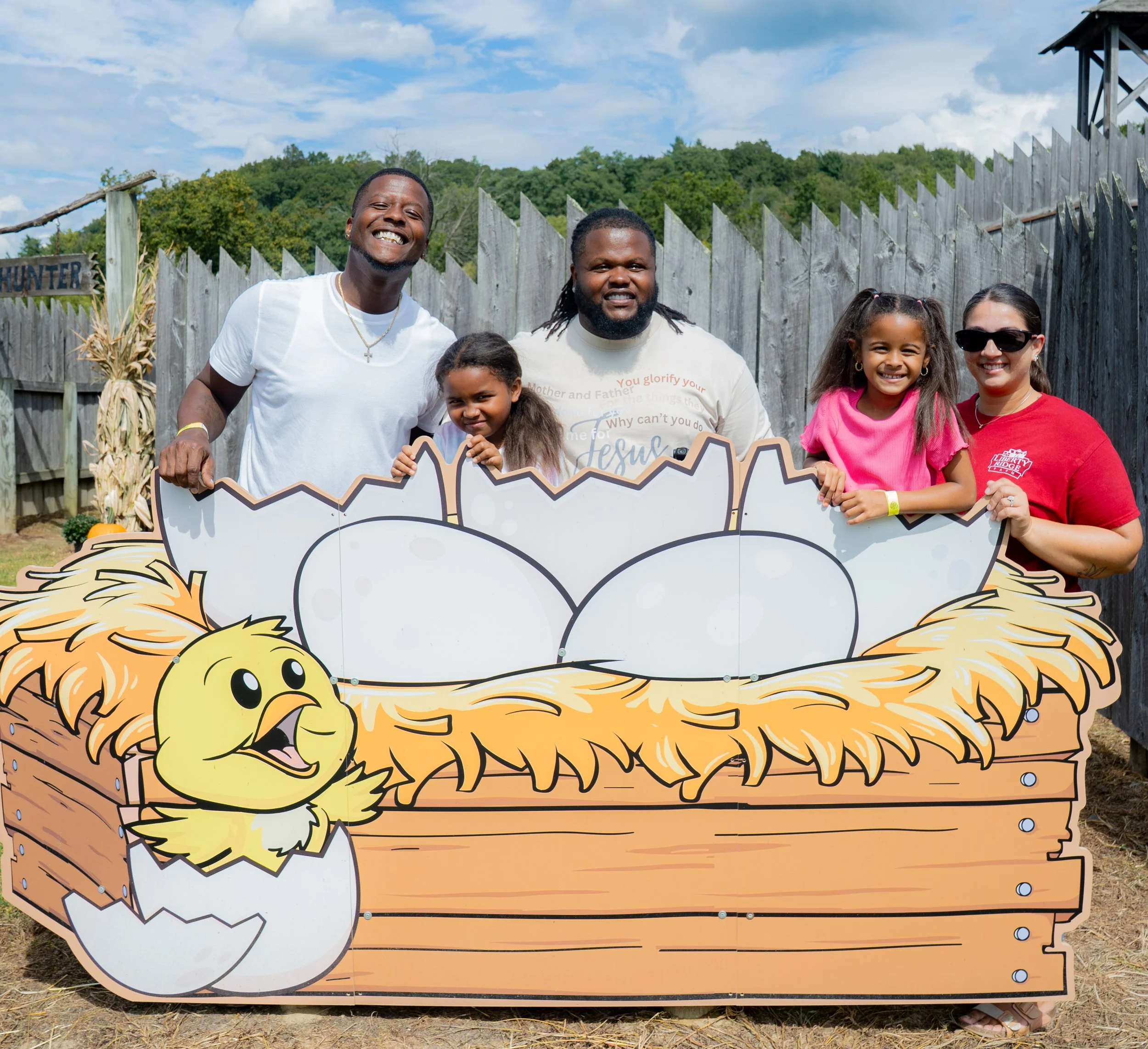 A group of people, including children and adults, standing behind a decorative cutout with a lion's face, outdoors against a wooden fence, trees, and a cloudy sky, smiling at the camera.