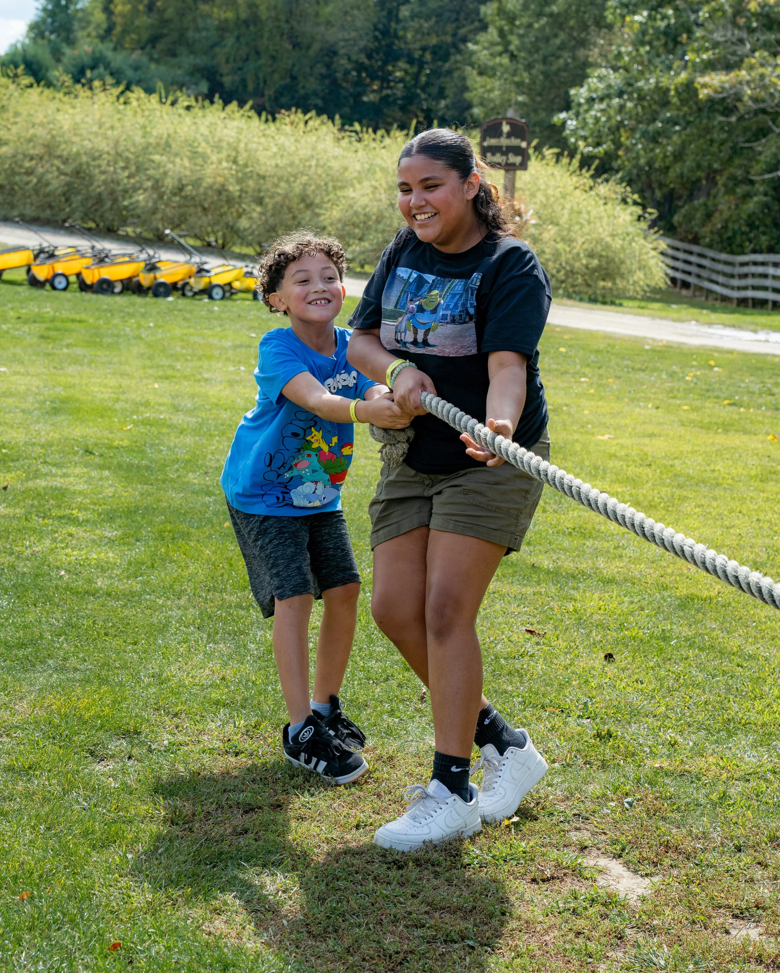 Two children, a girl and a boy, smiling and holding a rope in a grassy park during daytime. The girl has long dark hair, wearing a black T-shirt with a cartoon graphic, khaki shorts, white sneakers, and black socks. The boy has curly hair, wearing a blue T-shirt with a cartoon graphic and dark shorts, and black sneakers. In the background, there are yellow ride-on lawnmowers lined up and green trees.