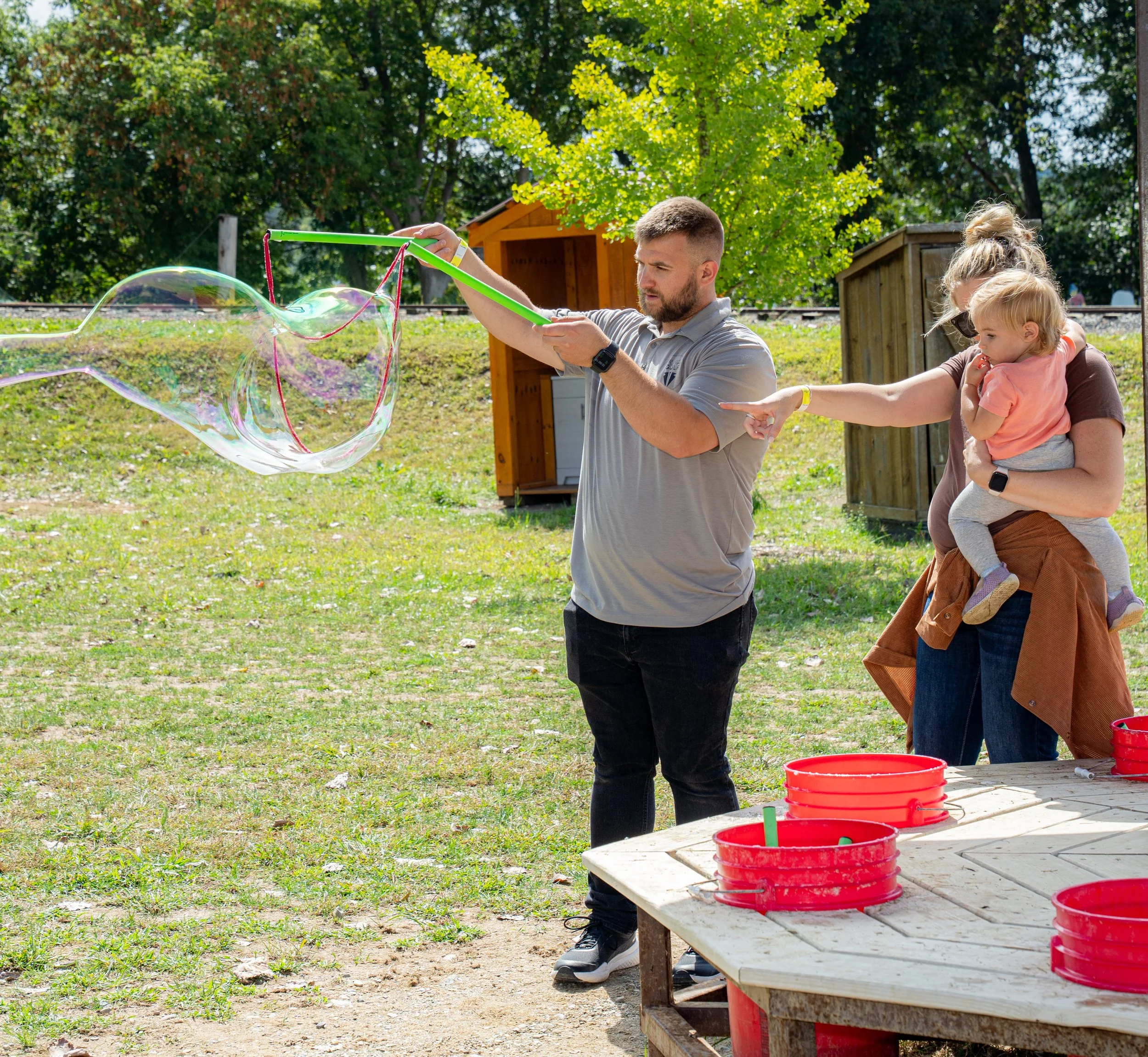 A man is creating large soap bubbles using a green bubble wand outdoors on a sunny day. Two women and a child are observing the bubbles, with trees and small wooden structures in the background. Red buckets are on a wooden table nearby.