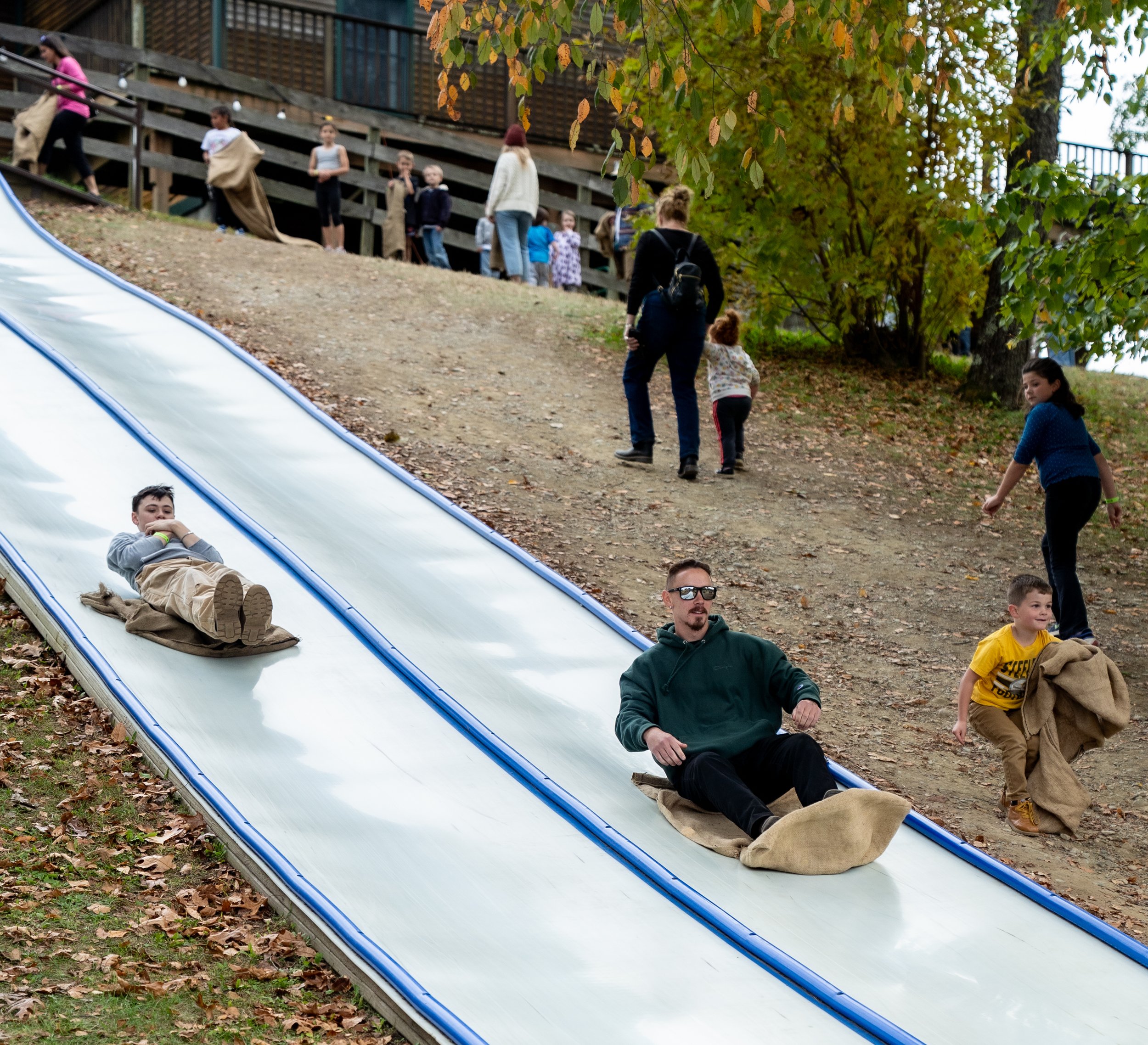 Children and adults playing on a large outdoor slide in a park during fall, with colorful autumn leaves on the ground and trees.