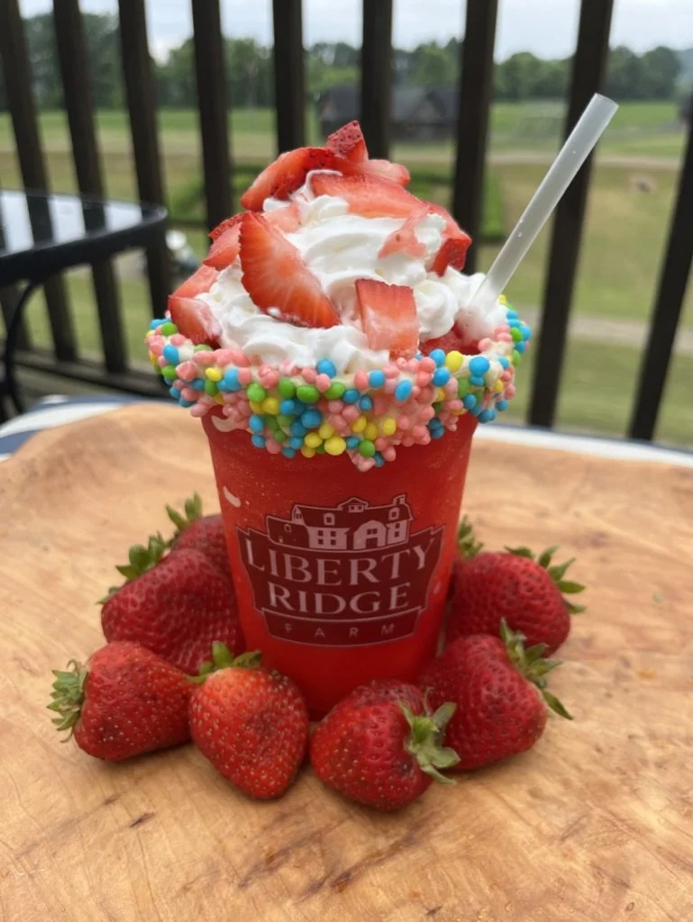 A strawberry-themed milkshake with whipped cream, strawberries on top, colorful sprinkles around the rim, and surrounded by fresh strawberries on a wooden plate with a blurred outdoor background.