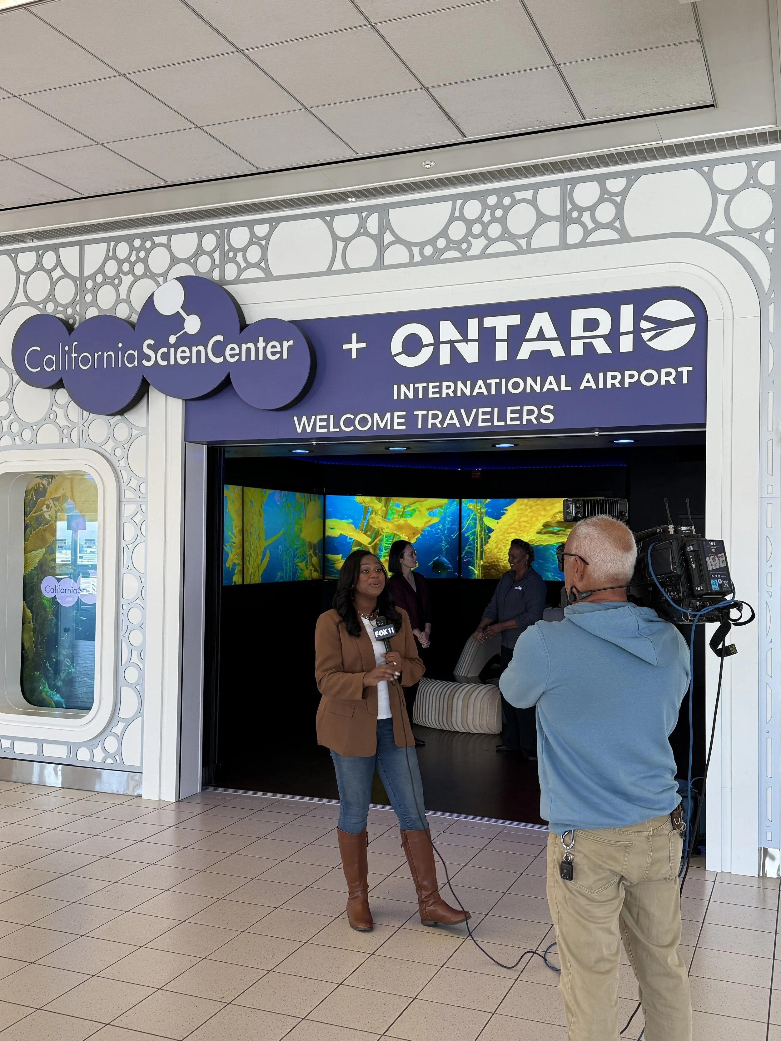 A Fox 11 Los Angeles reporter interviews representatives at the entrance to the California Science Center immersive ocean experience at Ontario International Airport, with kelp forest video panels glowing behind them.