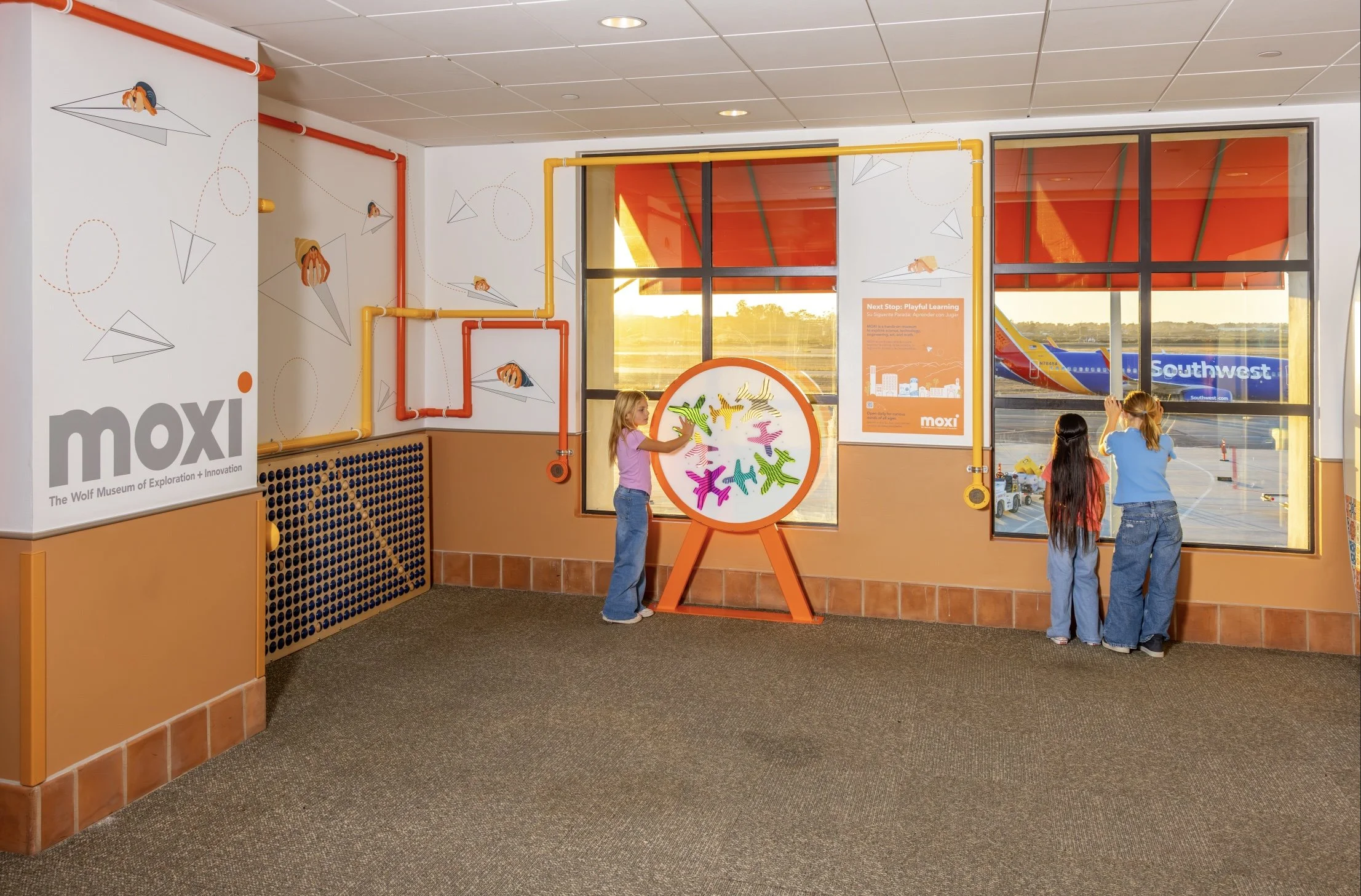 Children engage with the MOXI interactive exhibit at Santa Barbara Airport near Gate 2, featuring a colorful Airplane Color Mixing Wheel and tactile wall installations, with Southwest Airlines aircraft visible through the terminal windows.