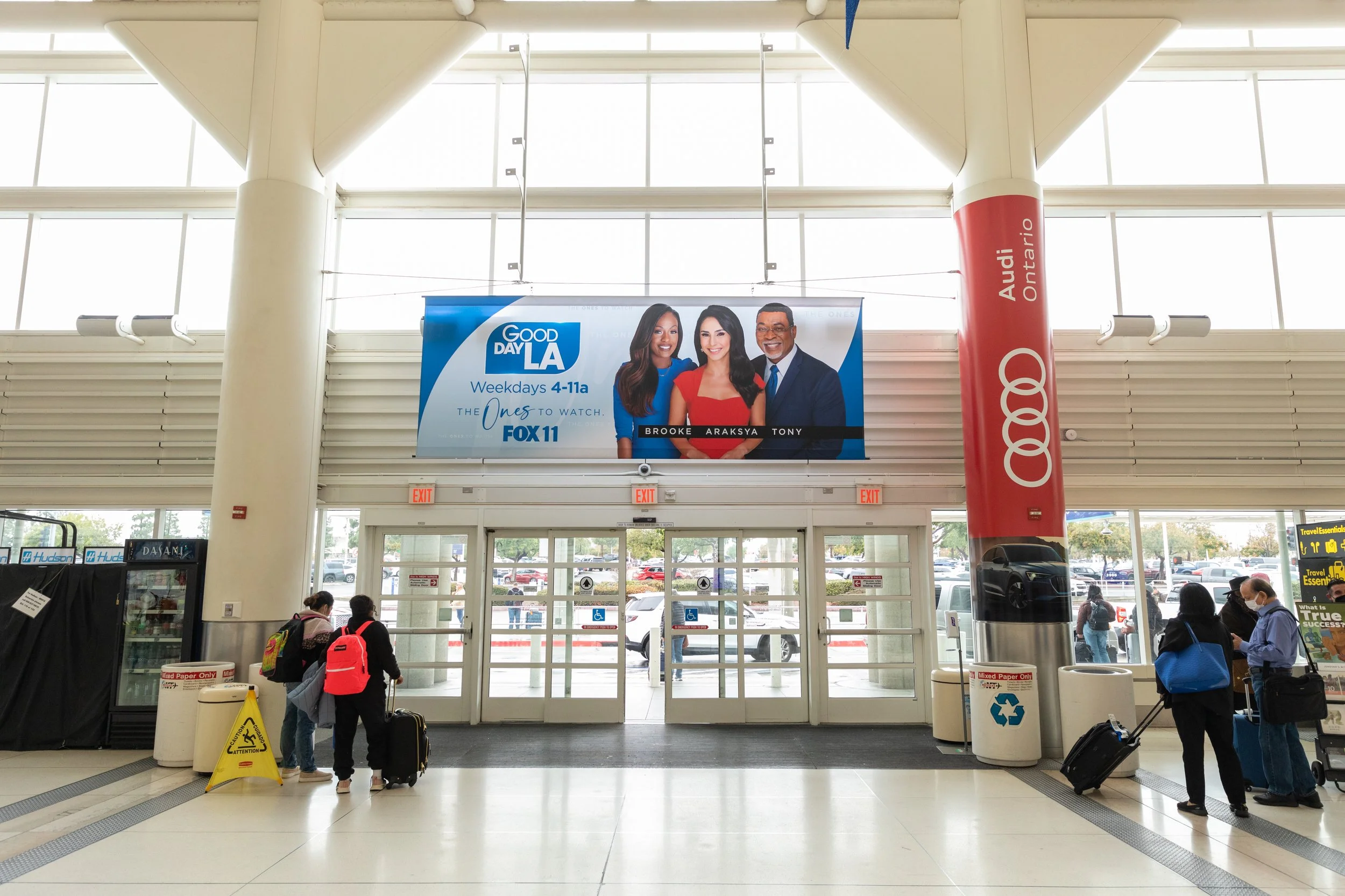 A large Good Day LA banner featuring hosts Brooke, Araksya, and Tony displayed above the exit doors in Ontario International Airport's terminal, with an Audi Ontario column wrap visible to the right.