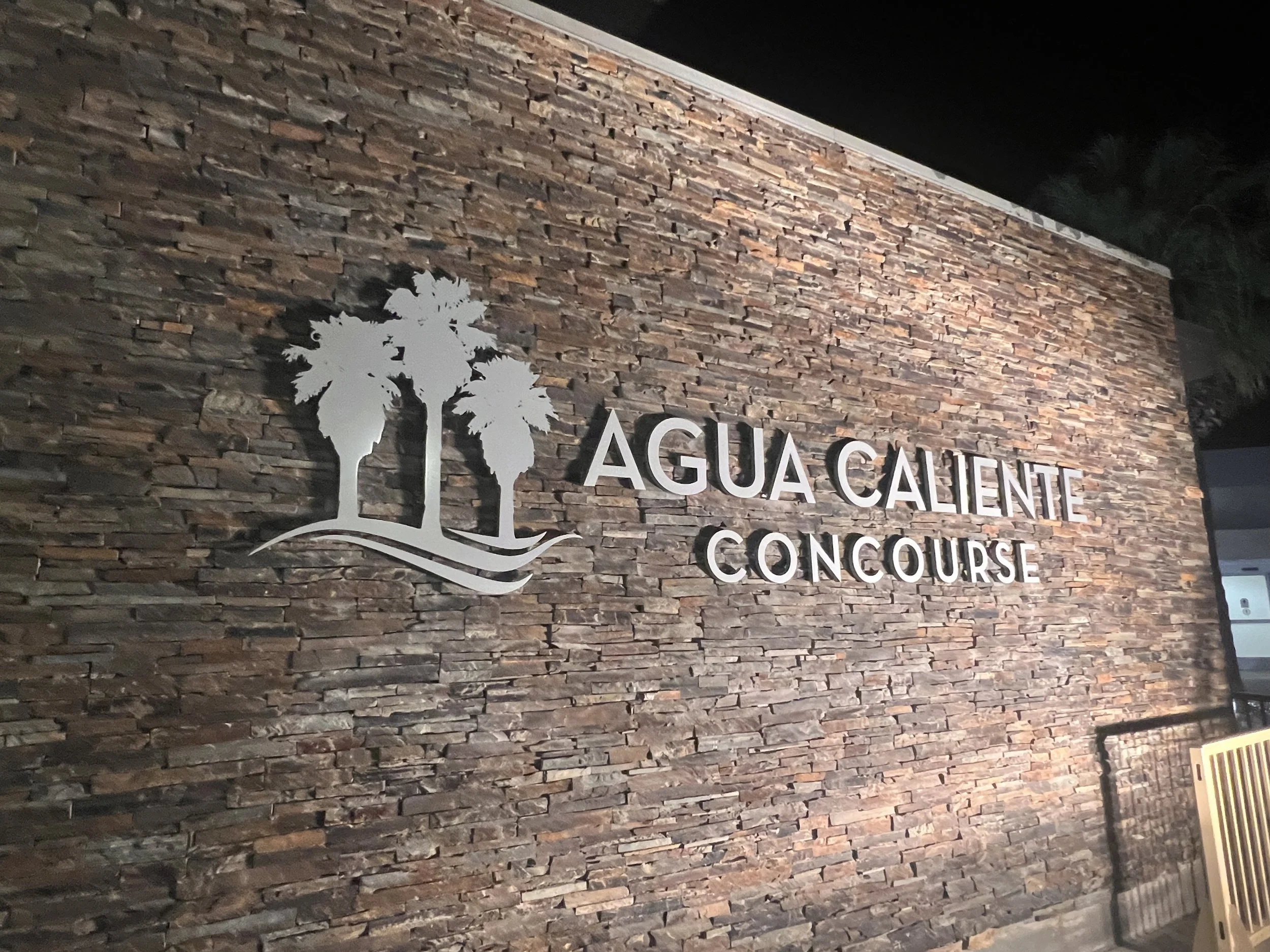 The Agua Caliente Concourse sign mounted on a textured stone wall at Palm Springs International Airport, featuring the Agua Caliente palm tree logo alongside the concourse name in brushed metal lettering.